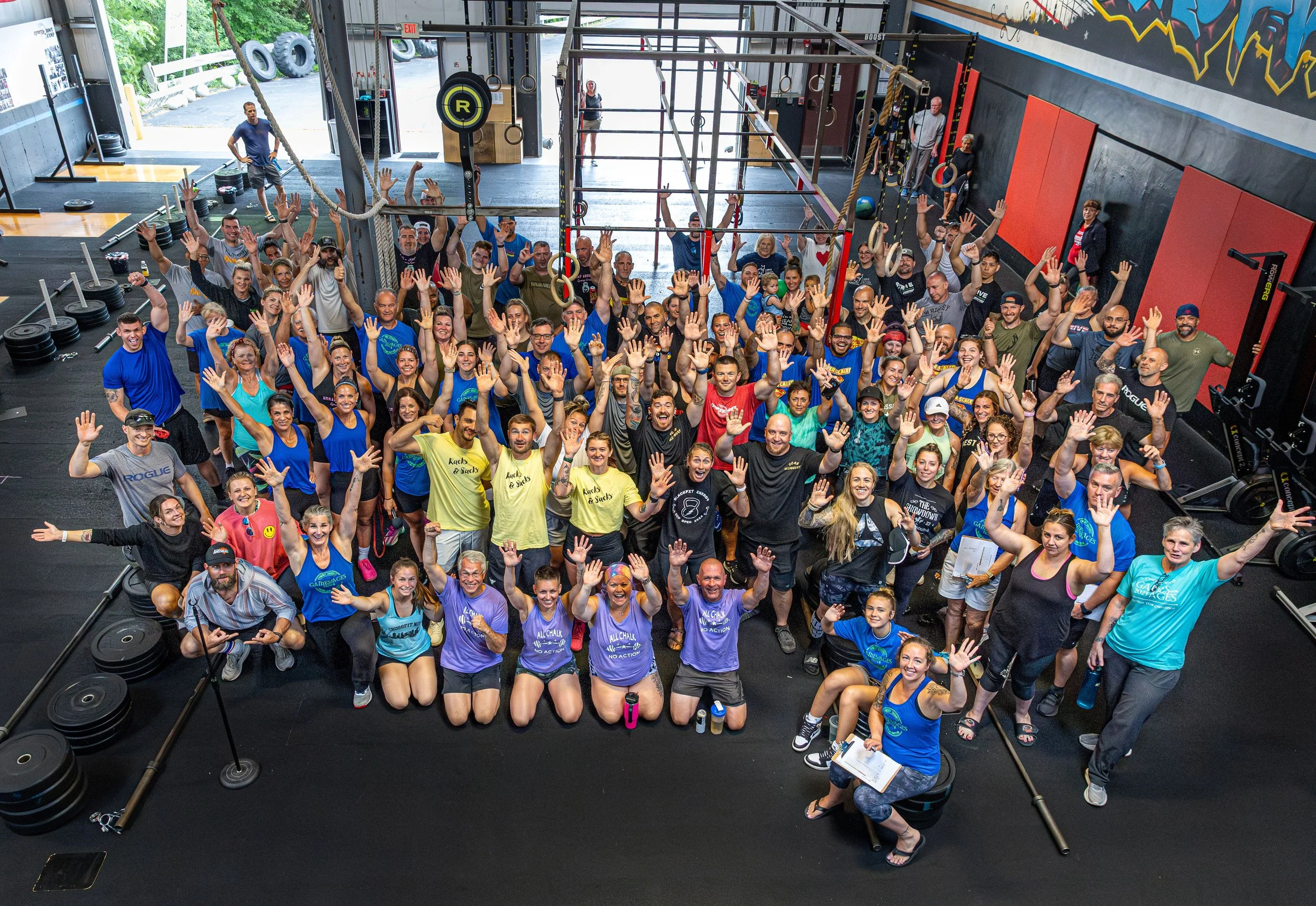 A large group of people gathered inside a gym, posing for a group photo with most people raising their hands. The gym has black flooring, fitness equipment like weights and rings, and colorful graffiti murals on the walls. The background shows open garage doors leading outside.