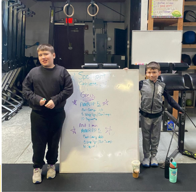 Two boys standing next to a whiteboard with workout instructions in a gym. The boy on the left has short hair and is wearing a dark sweatshirt and pants. The boy on the right has short hair and is wearing a gray and black jacket with black pants. The gym has workout equipment like pull-up rings and kettlebells in the background, and there is a water bottle and a trash cup in front of the whiteboard.