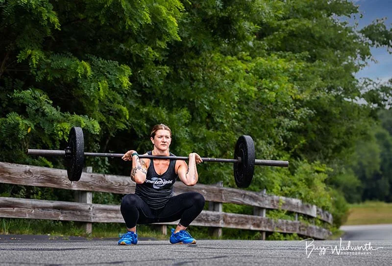 A woman is performing a squat exercise outdoors on a paved path, holding a barbell with weights across her shoulders, with green trees and a wooden fence in the background.