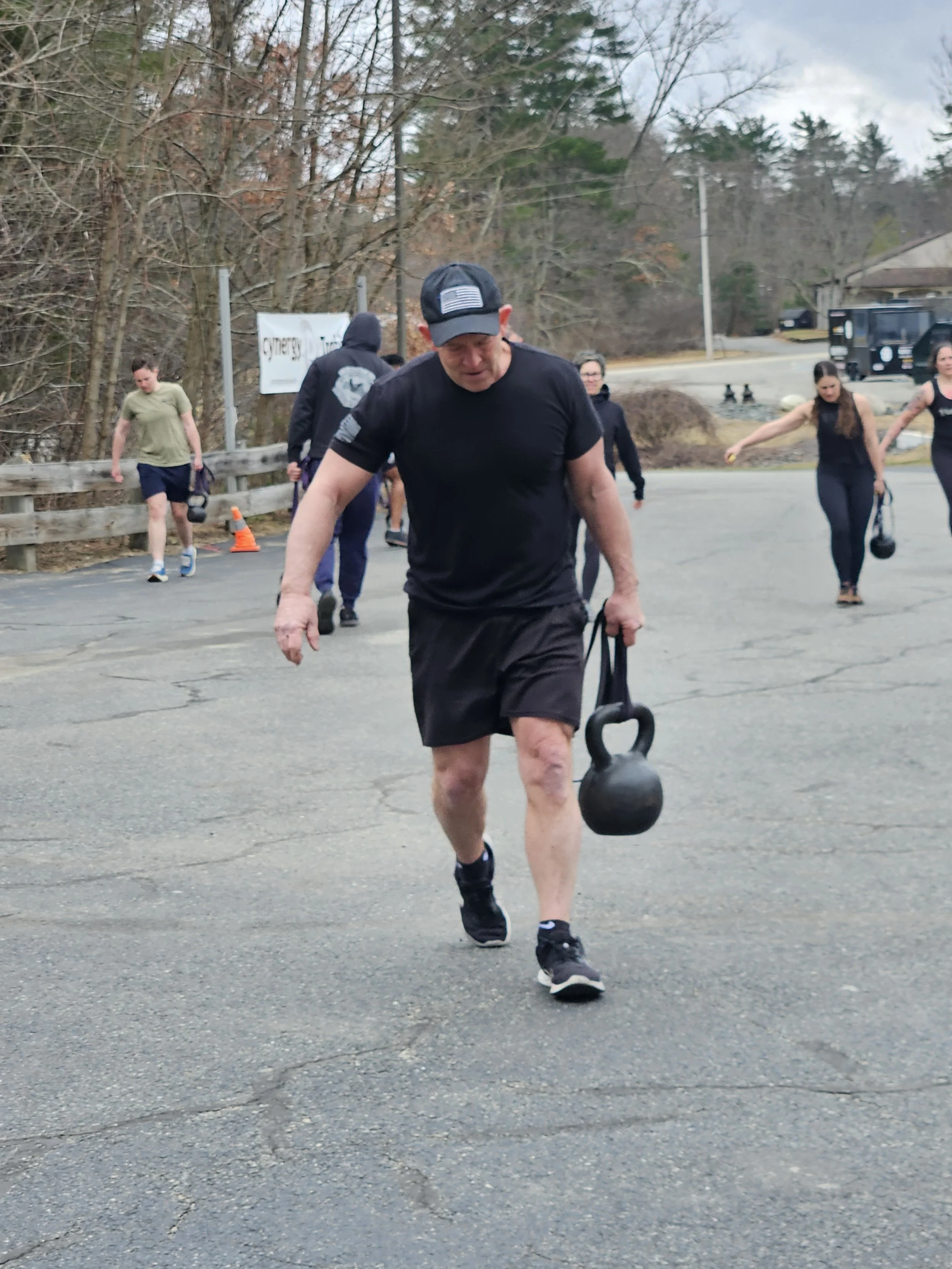Man walking on pavement carrying a kettlebell with several people exercising in the background outdoors.