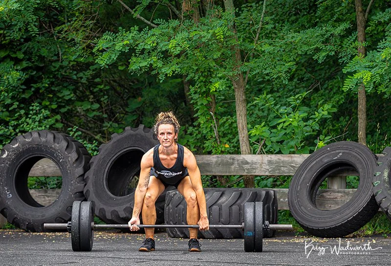 A woman lifting a barbell outdoors with large tires in the background.