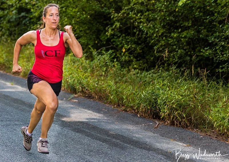 Woman running outdoors on a paved road, wearing a red tank top and black shorts with purple running shoes, surrounded by green foliage.