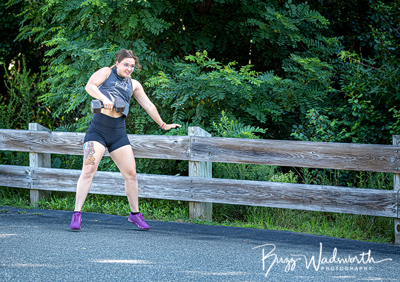 Woman exercising outdoors on a paved trail, holding a dumbbell, wearing workout clothes and purple shoes, with a wooden fence and green foliage in the background.