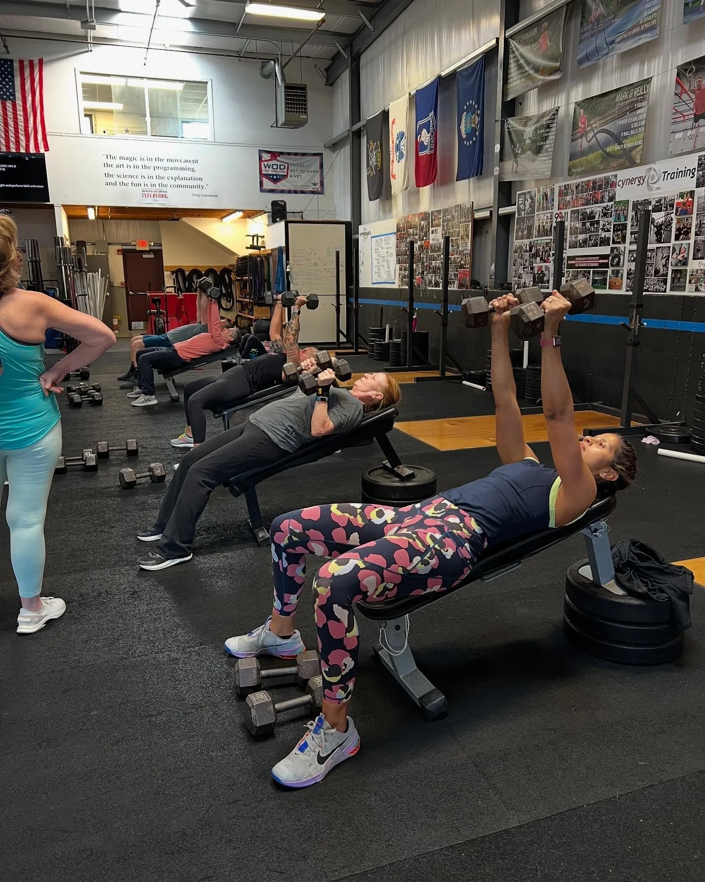 People lifting dumbbells on workout benches in a gym, with a trainer supervising. The gym has motivational posters, flags, and photographs on the walls.