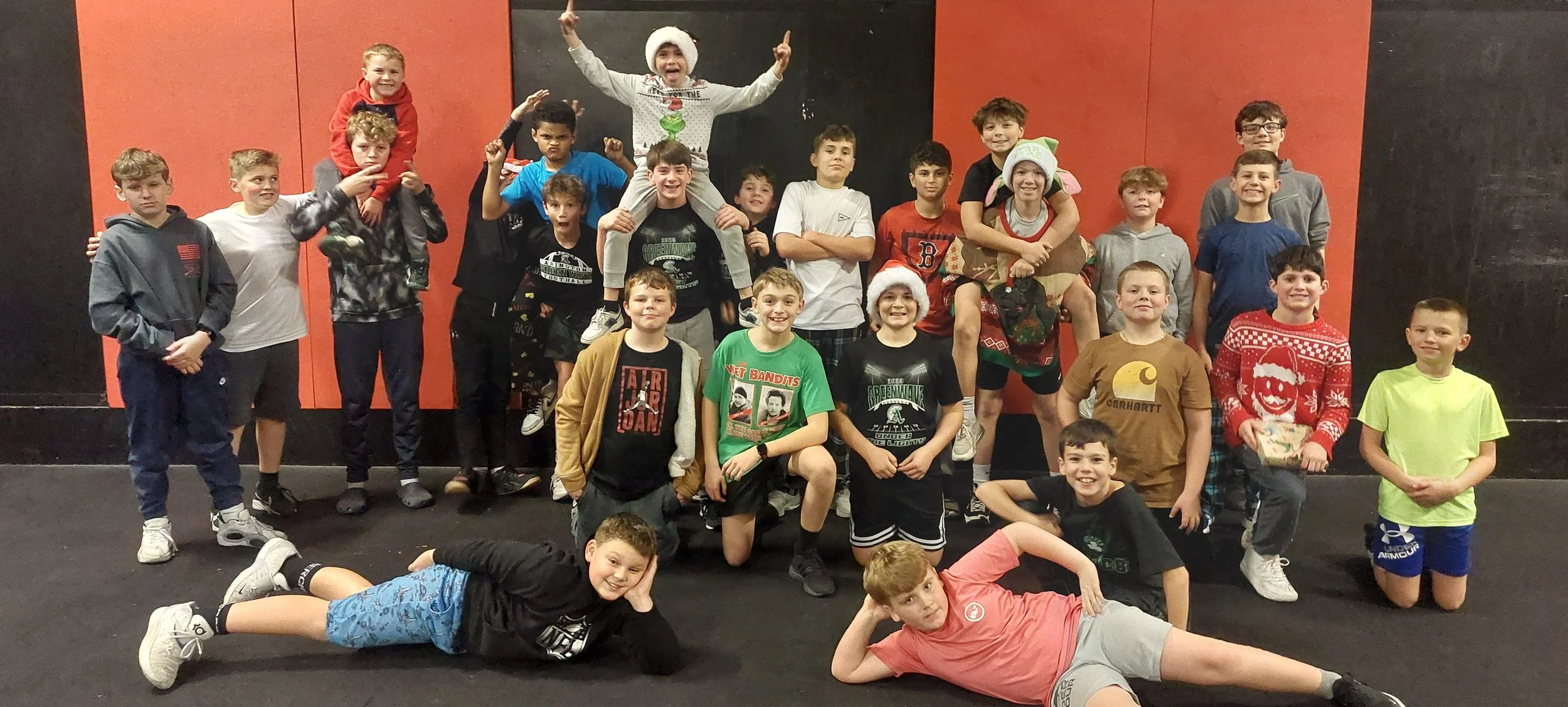 A group of boys and young teens, some wearing Christmas hats and sweaters, pose together in a gym or recreational center, with some standing, some crouching, and a few lying on the floor, celebrating during a festive or holiday event.