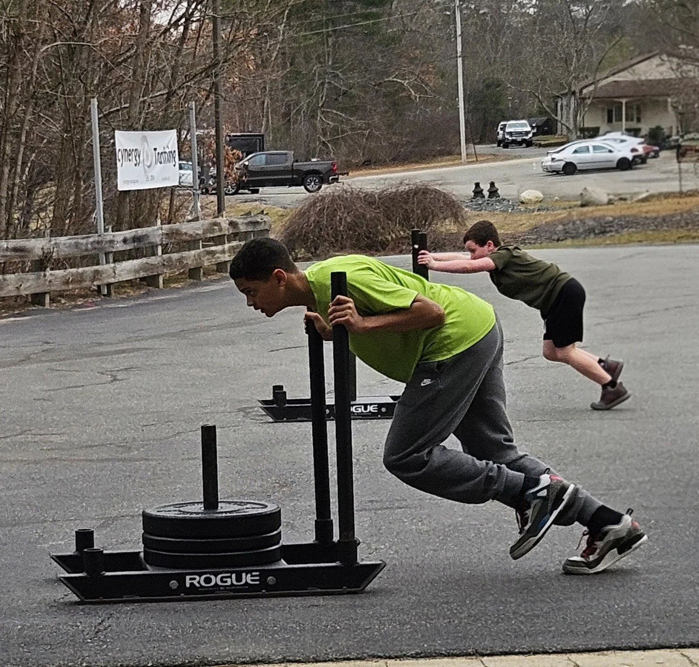 Two boys pushing a weighted sled outdoors in a parking lot. The boy in front is wearing a green shirt and black pants, and the boy in the back is wearing a dark green shirt and shorts, both appearing to be pushing the sled together.