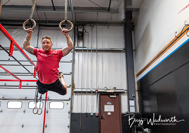 Man performing gymnastic rings exercise in gym with black and white walls, red railing, and overhead garage-style doors.