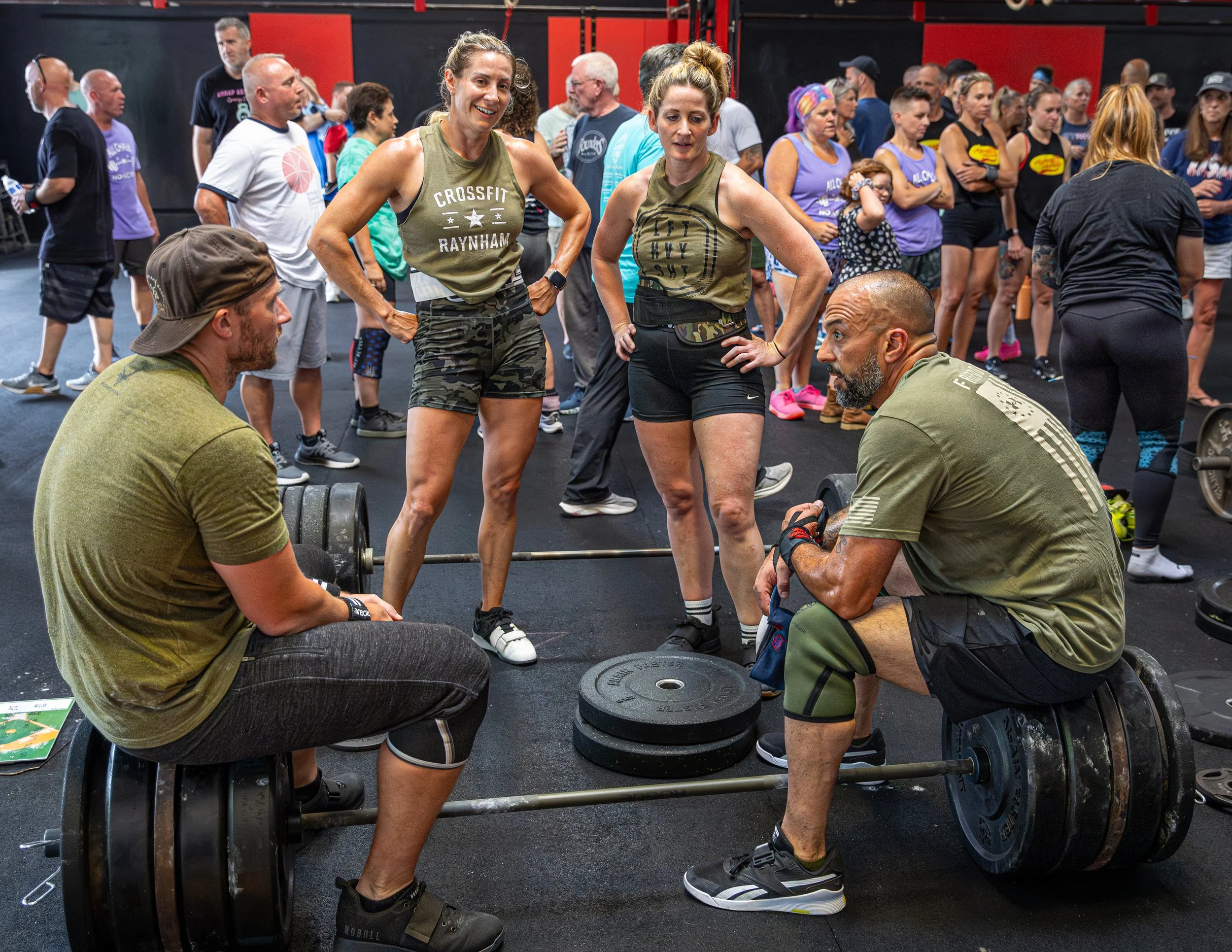 People at a CrossFit gym participating in a weightlifting workout, with some spotting others lifting weights and a large crowd watching.