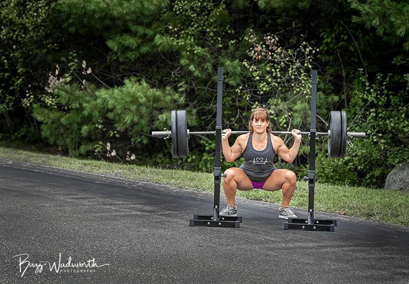 A woman lifting a barbell in an outdoor setting with trees in the background.