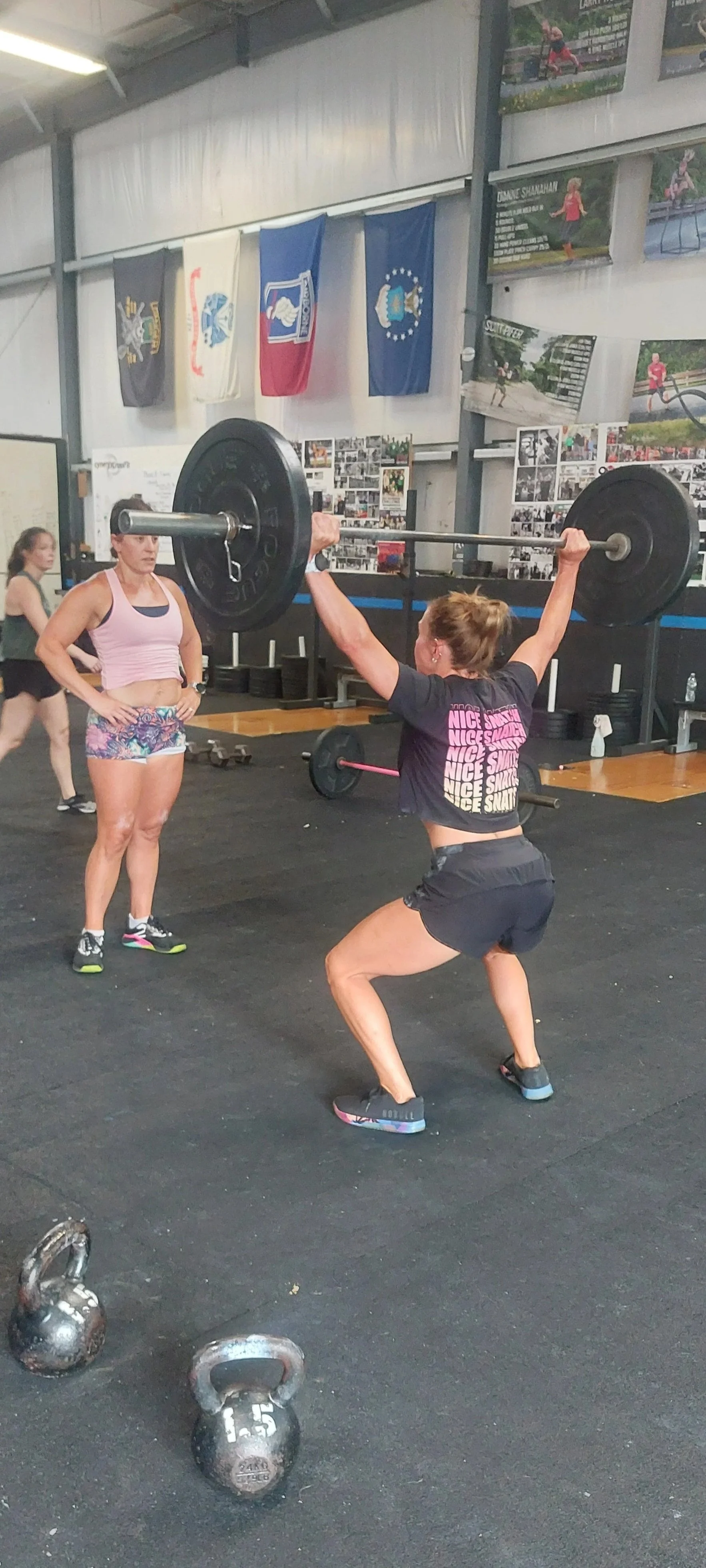 A woman is performing an overhead barbell squat in a gym with black rubber flooring, while two kettlebells are on the ground nearby. In the background, other women are working out and flags and posters are hanging on the wall.