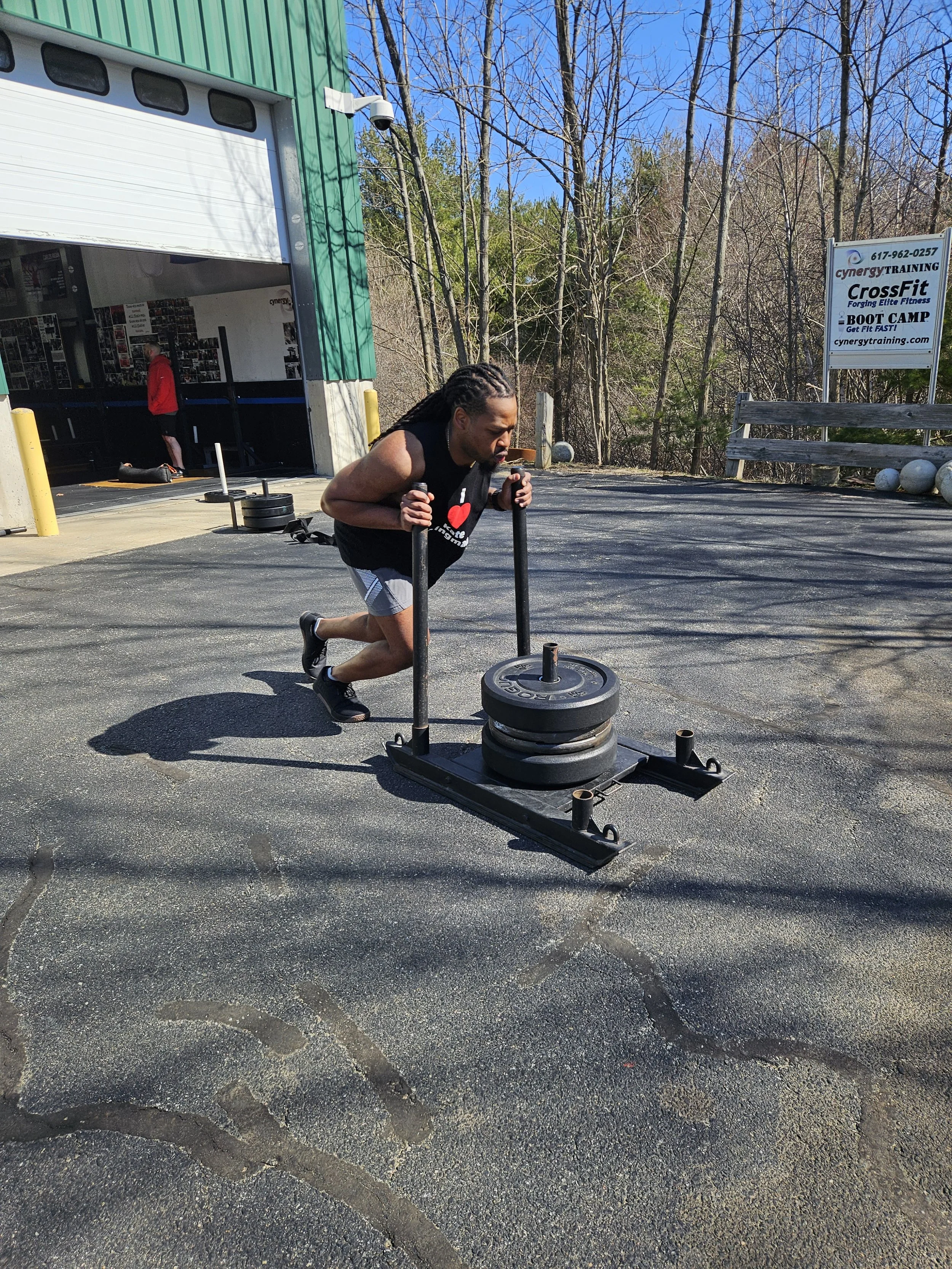 A man with dreadlocks wearing a black sleeveless shirt is pushing a sled loaded with weight plates during outdoor workout outside a gym. There is a person in a red jacket inside the gym, and a sign for CrossFit Boot Camp is visible in the background amidst leafless trees.
