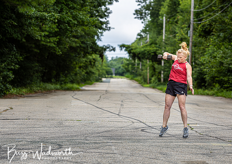 Woman in red tank top and black shorts lifting dumbbells while standing on an empty road surrounded by green trees.