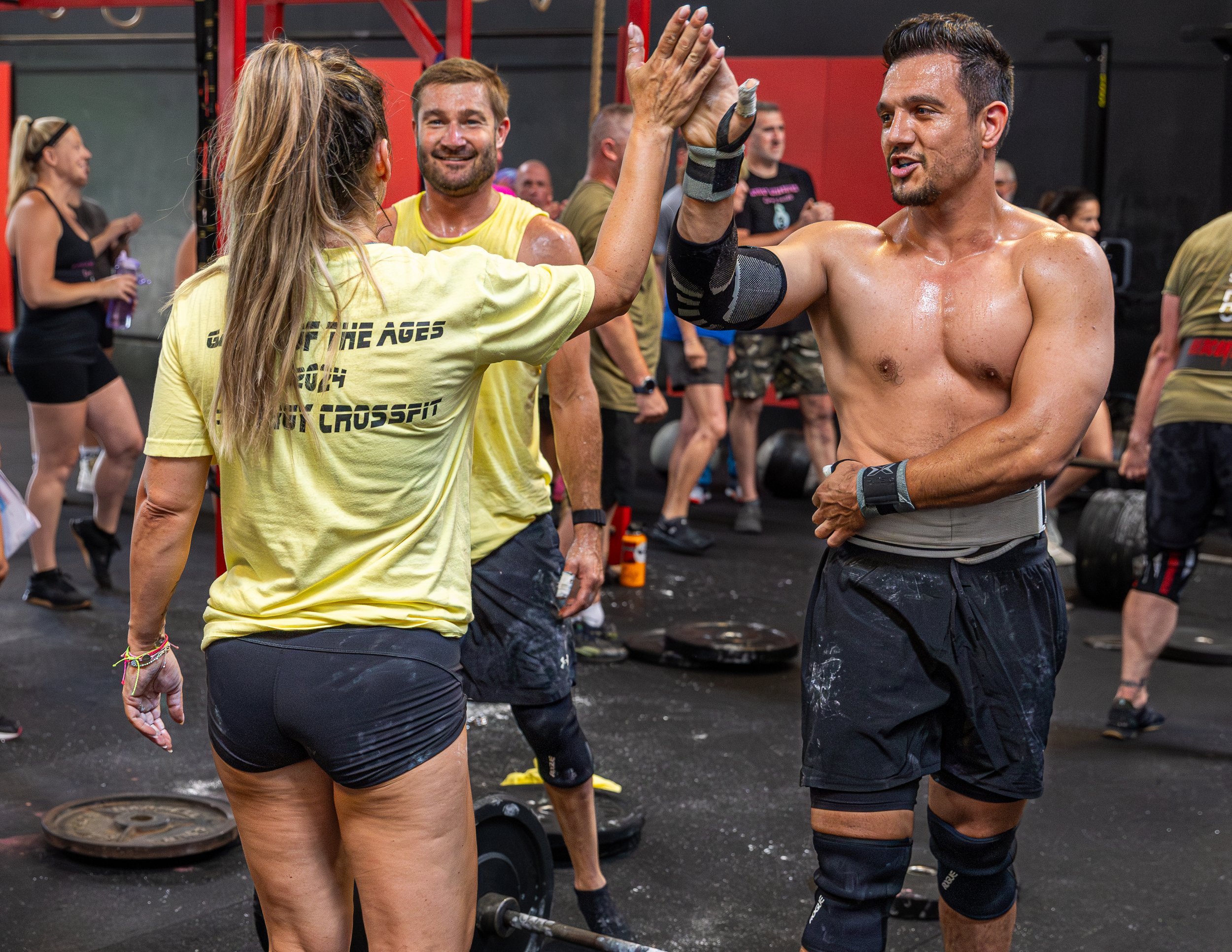 Two people high-fiving at the gym, one wearing a yellow shirt and the other shirtless with black shorts, surrounded by other gym-goers and workout equipment.