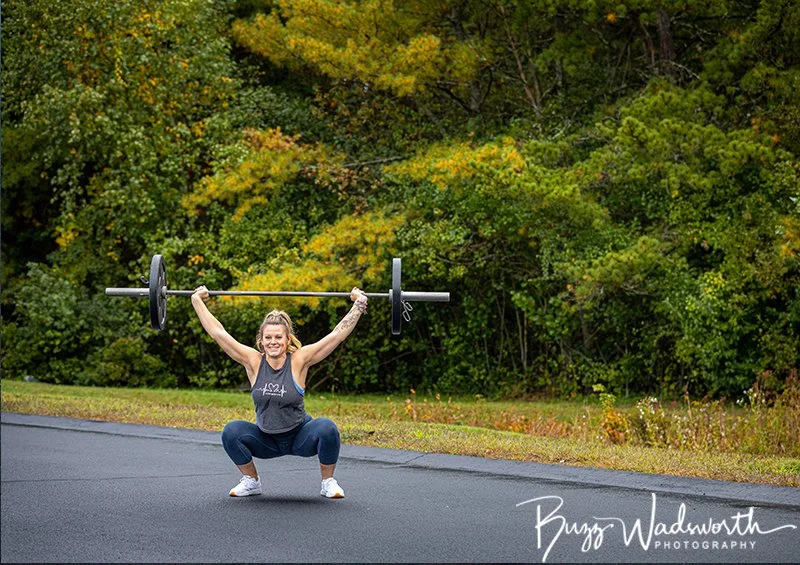 A woman lifting a barbell in a squat position outdoors on a paved road, with a background of lush green trees.