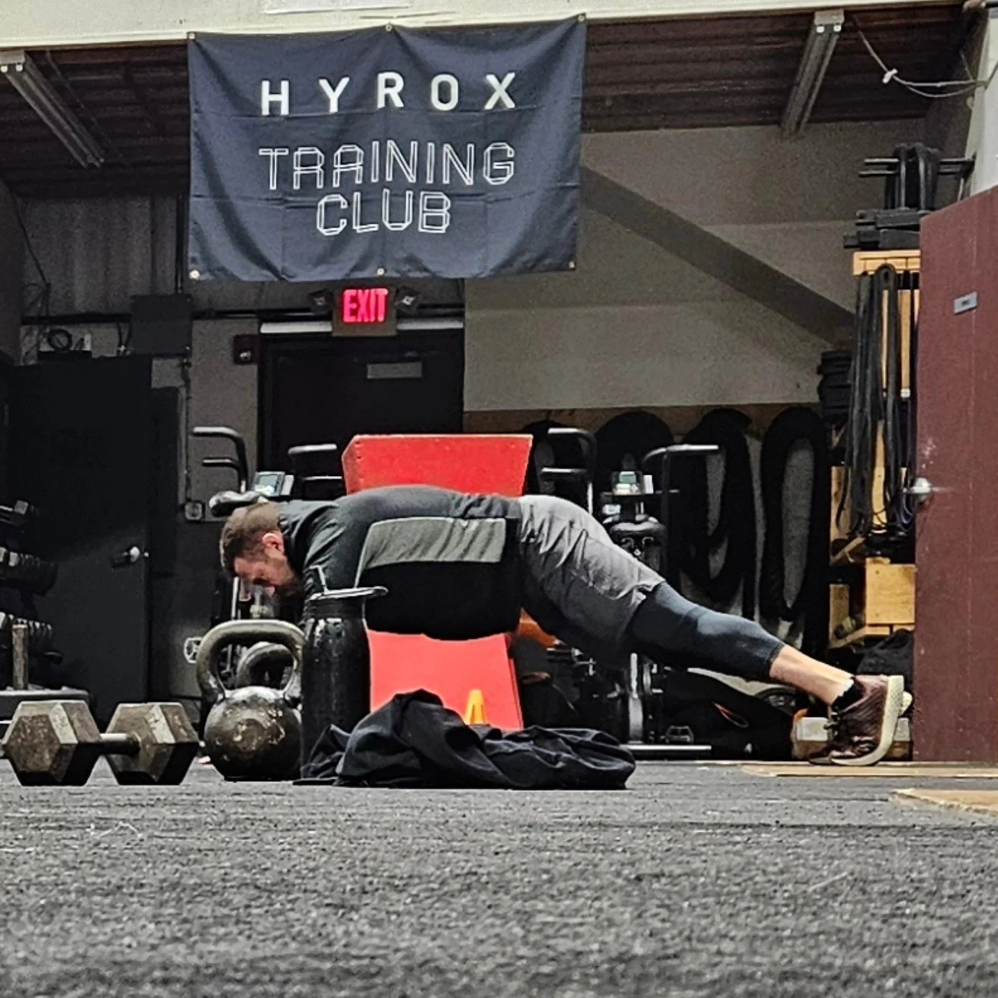 A man performing a plank exercise on a workout mat inside a gym. The gym has a sign that reads 'HYROX TRAINING CLUB' and various weights and equipment around.