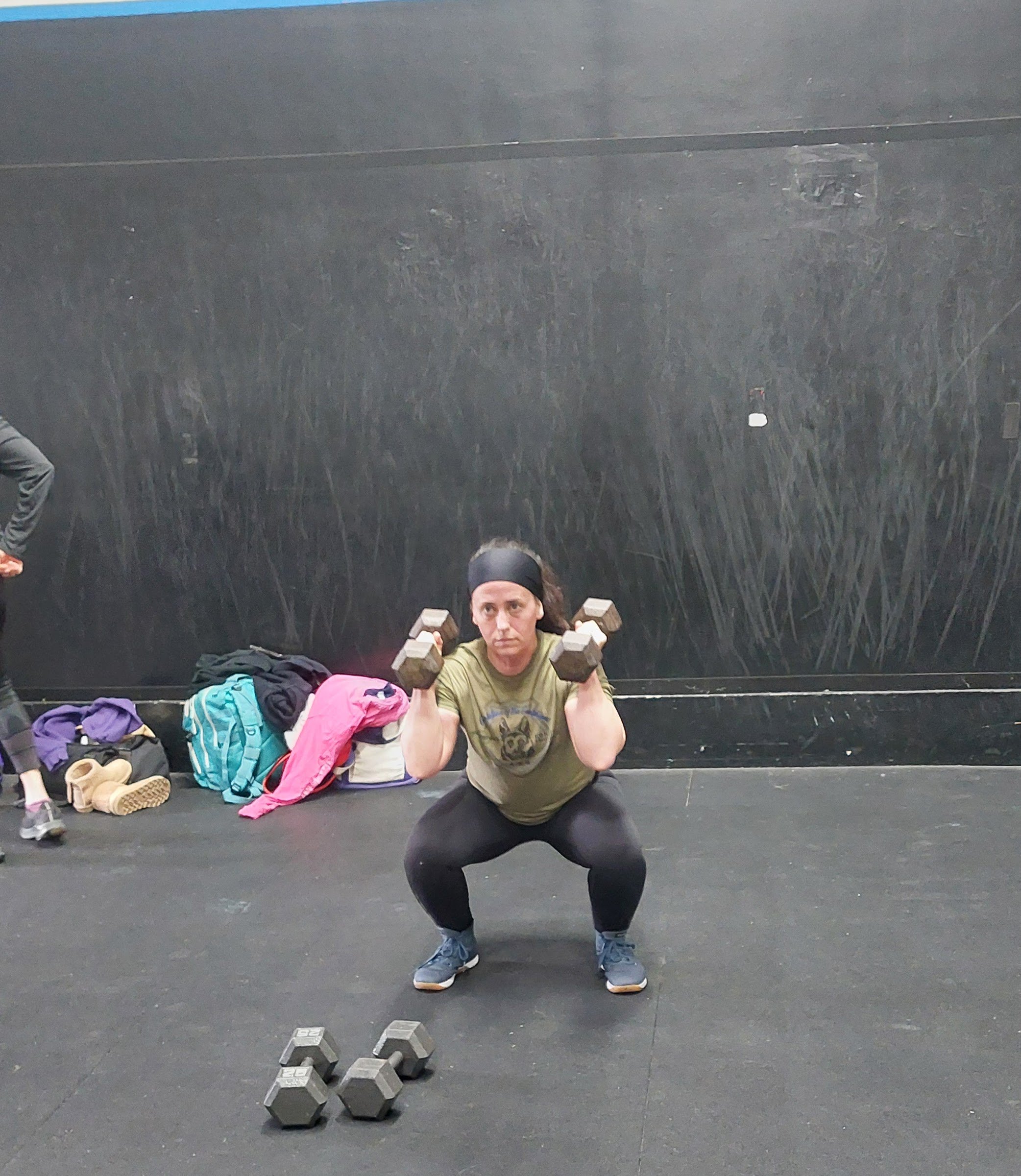 A person wearing a black headband, green shirt, and black pants performing a squat with dumbbells in a gym. There are colored backpacks, clothing, water bottles, and shoes on the floor, and a group of dumbbells is in the foreground.
