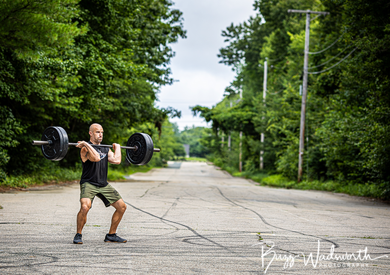 A man lifting a barbell with weights outdoors on a cracked road surrounded by green trees.