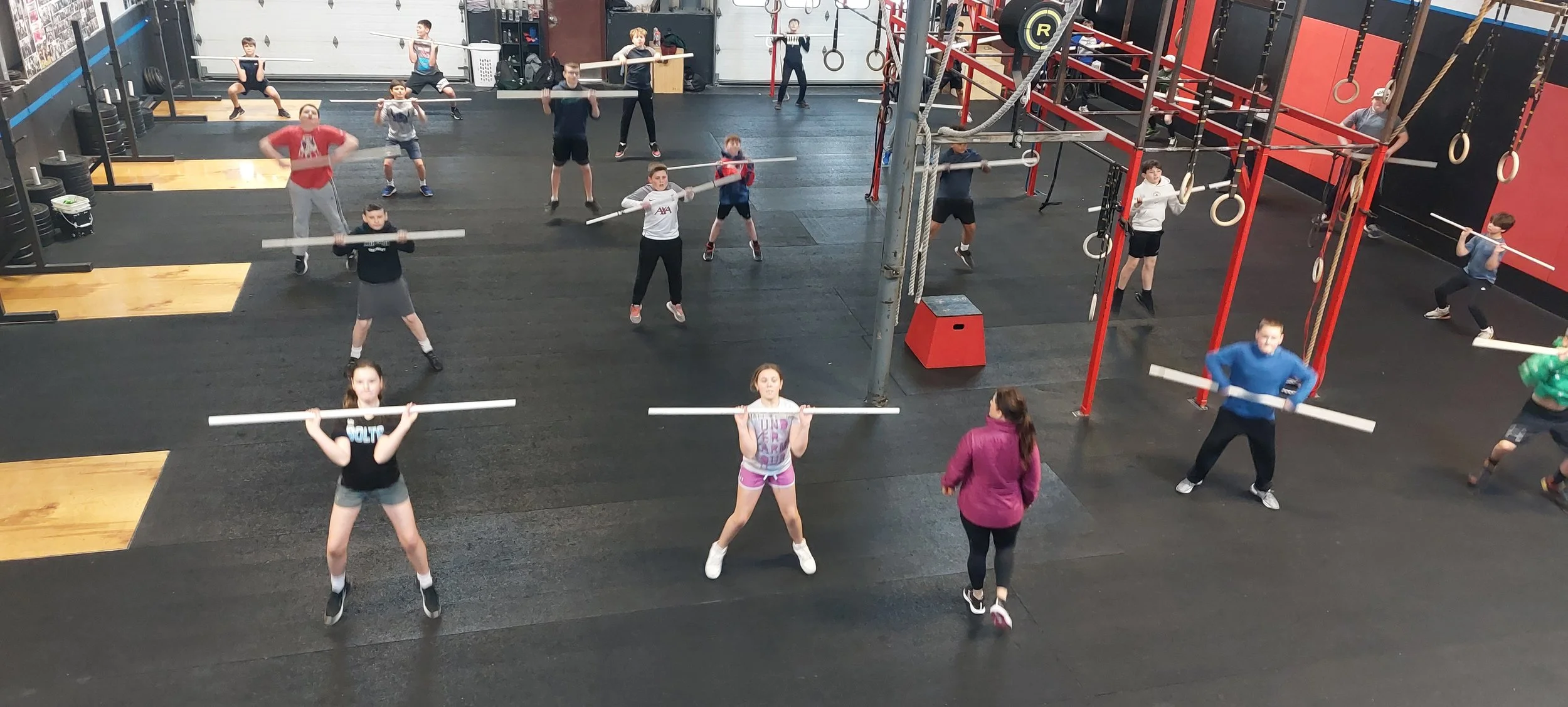 Group of children and teenagers in a gym, lifting and holding long white bars during a fitness class, supervised by an instructor in a pink jacket.