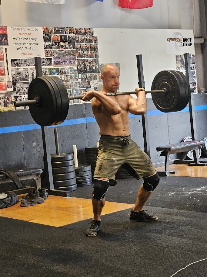 A man lifting a heavy barbell with weights in a gym, wearing shorts, knee braces, and black shoes, positioned in a squat stance.