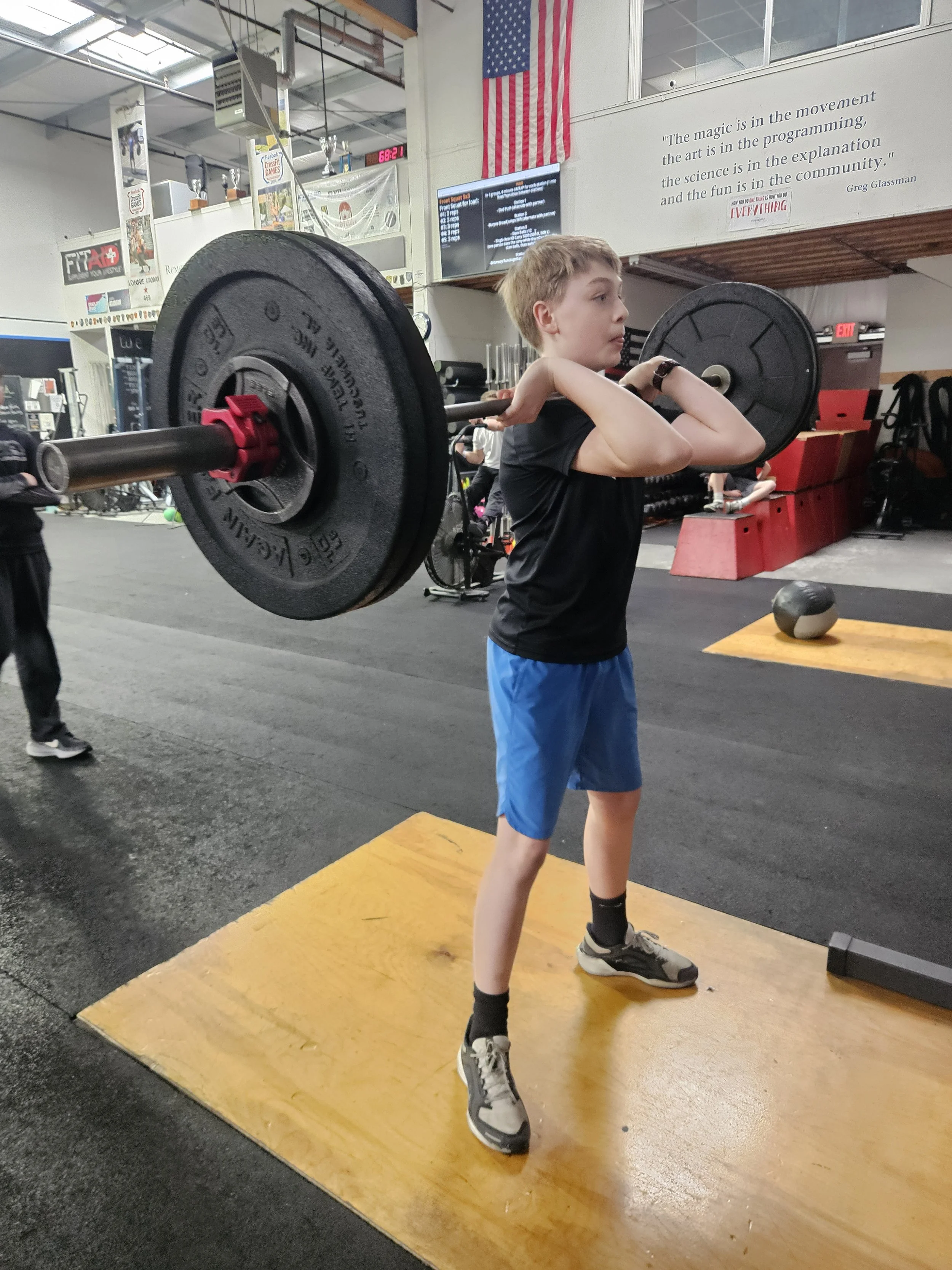 A young boy lifting a barbell with weights at a gym, standing on a yellow platform.