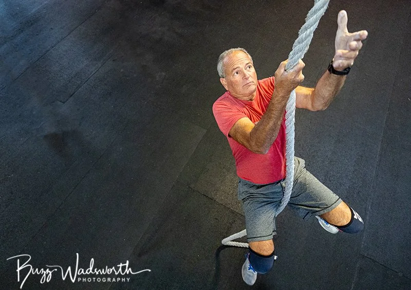 An older man in a red shirt and gray shorts climbing a rope in a gym, looking up.
