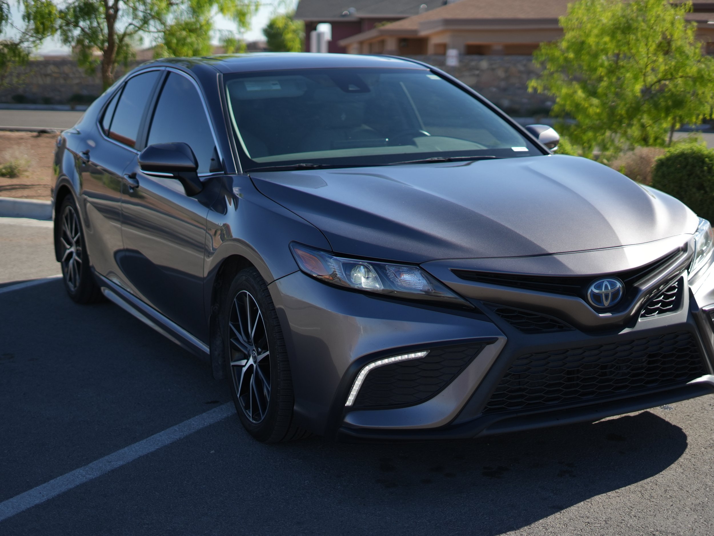 A dark gray Toyota sedan parked in an outdoor parking lot with yellow designated parking lines, green trees, and residential buildings in the background.