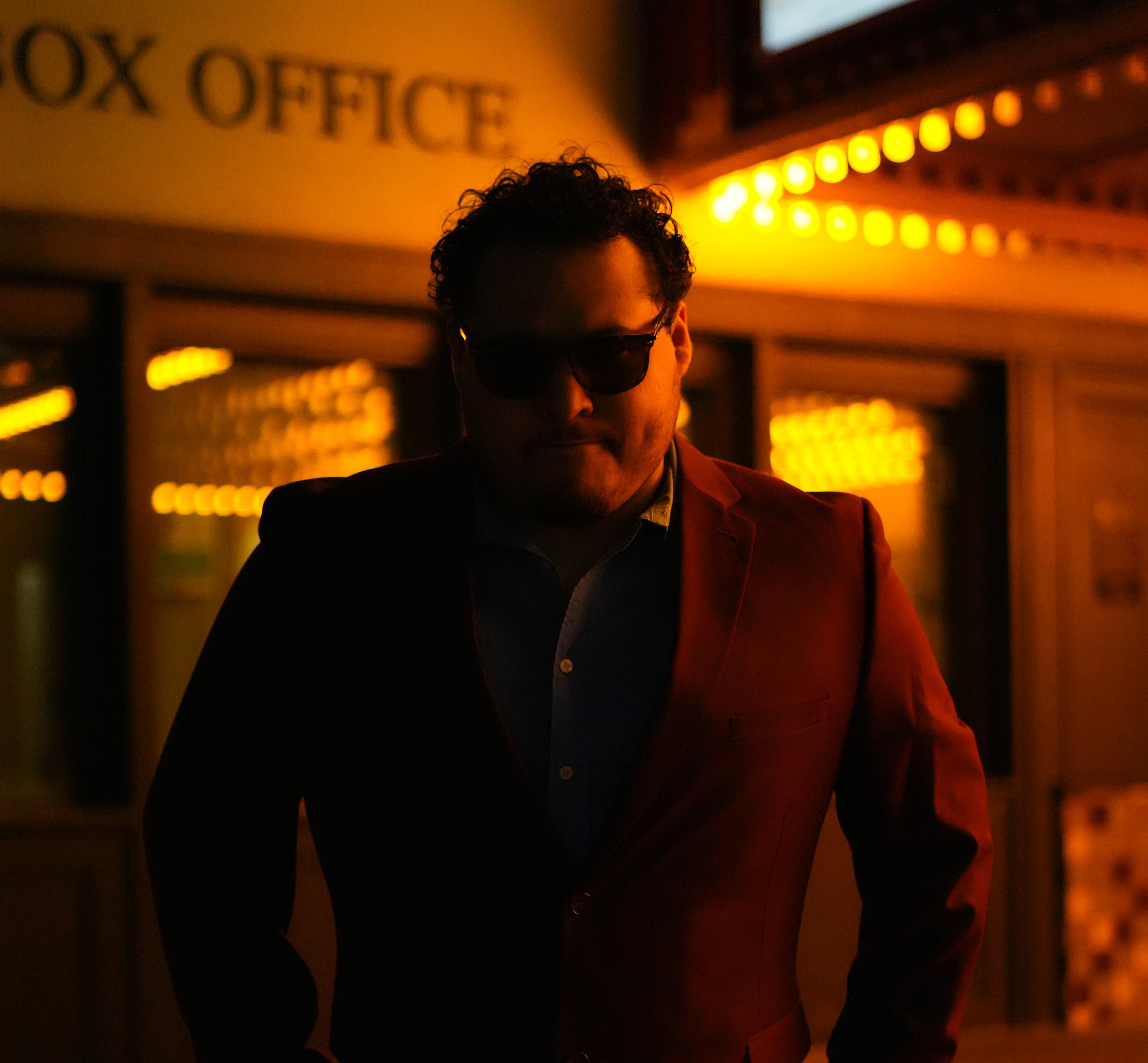 A person with curly hair wearing sunglasses and a suit, standing in front of a glowing maroon and yellow background with the words 'BOX OFFICE' visible on a sign.