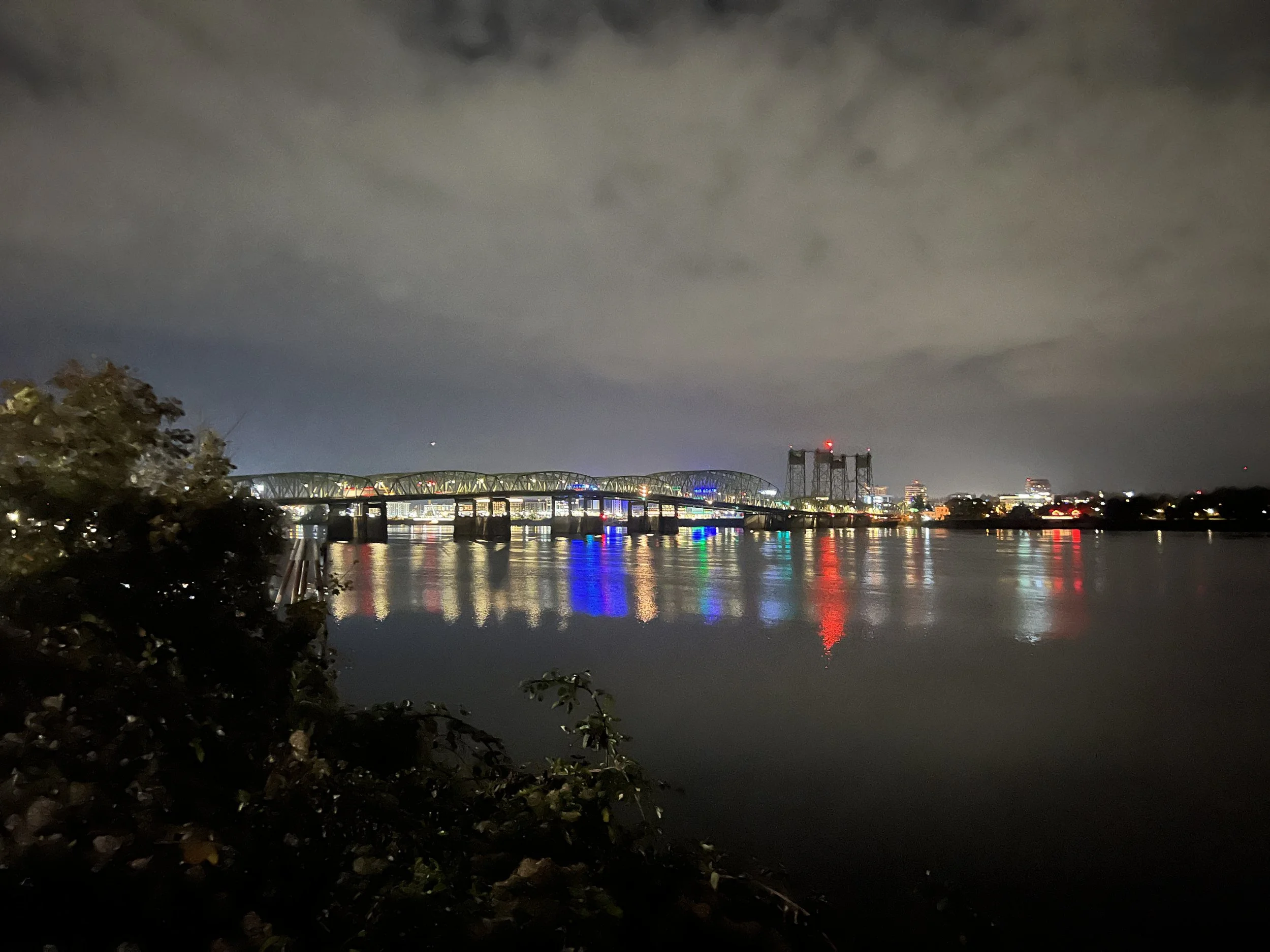 Nighttime cityscape with a river reflecting colorful lights, a bridge, buildings with illuminated windows, and a dark sky with clouds and a small bright spot.