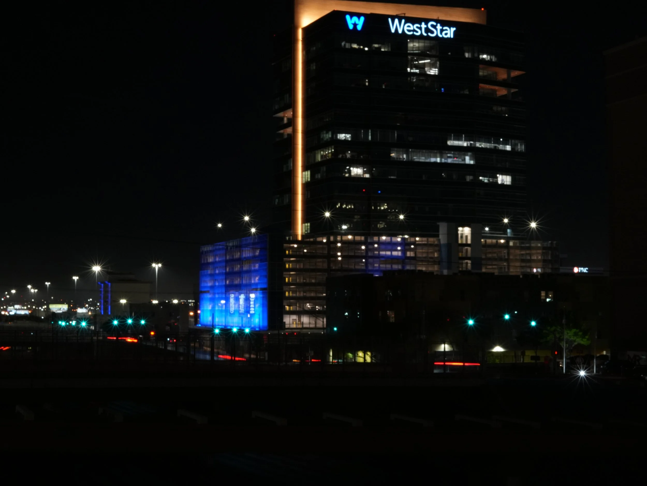 Nighttime view of a tall building with the WestStar logo illuminated at the top. The building features glass windows with interior lights on, and the lower part of the building is accentuated with blue lighting. In the foreground, there are streetlights and blurred car lights.