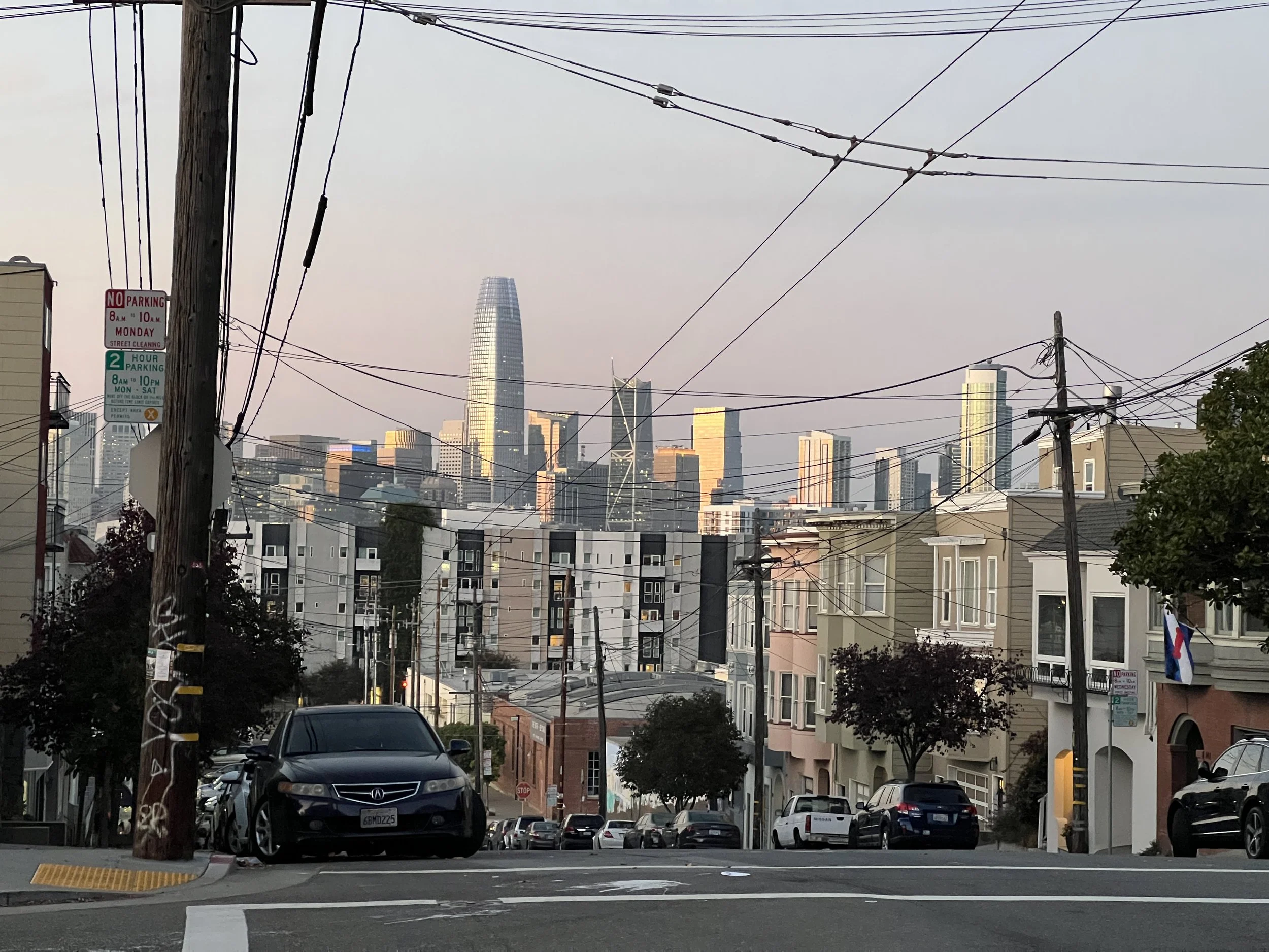 Cityscape view with buildings, power lines, and a parking lot, during dusk in San Francisco with the Salesforce Tower visible in the distance.