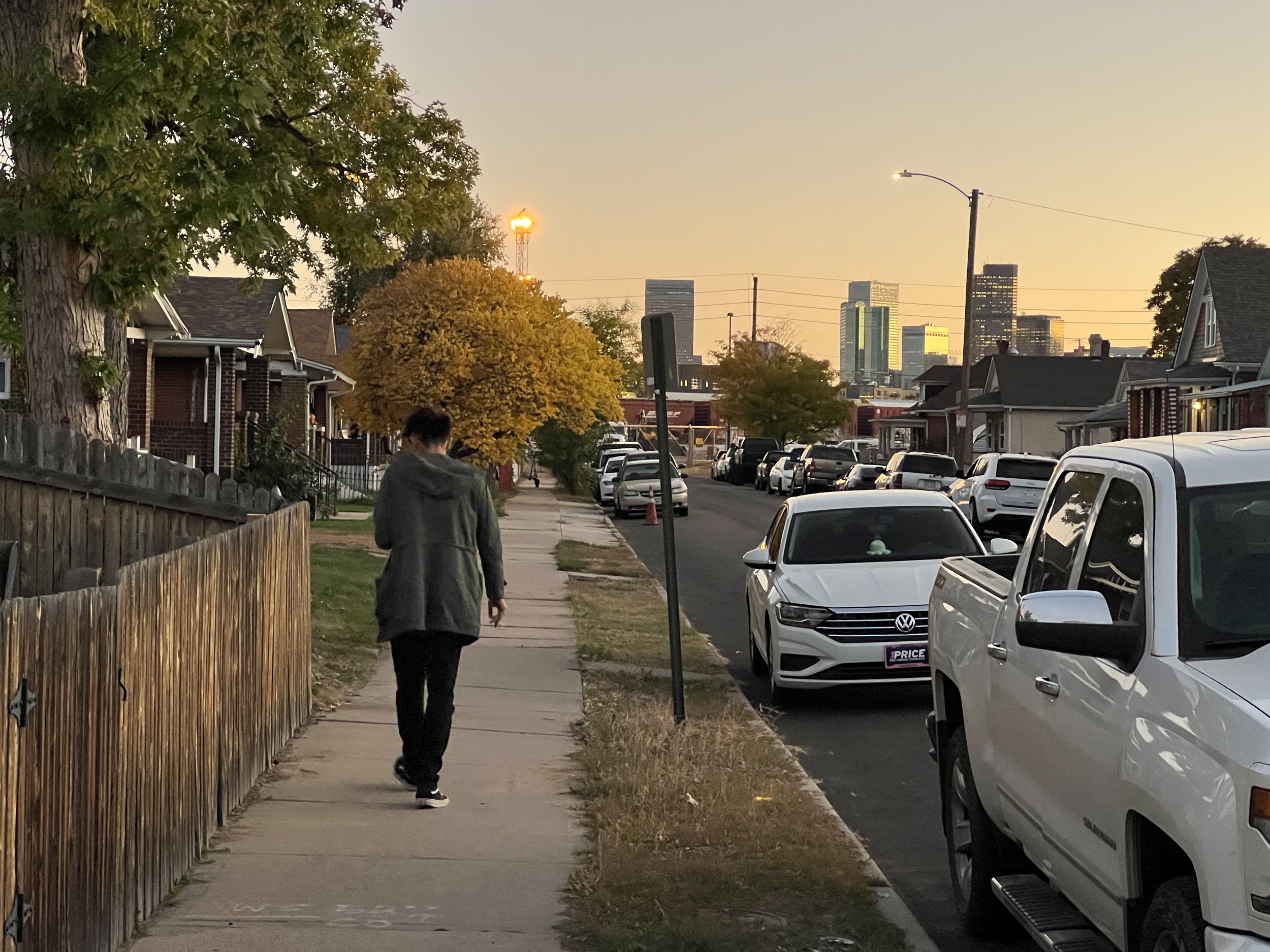 A residential street with parked cars along the curb, a person walking on the sidewalk, trees with autumn foliage, houses with brick and siding exteriors, and a city skyline in the distance during sunset.