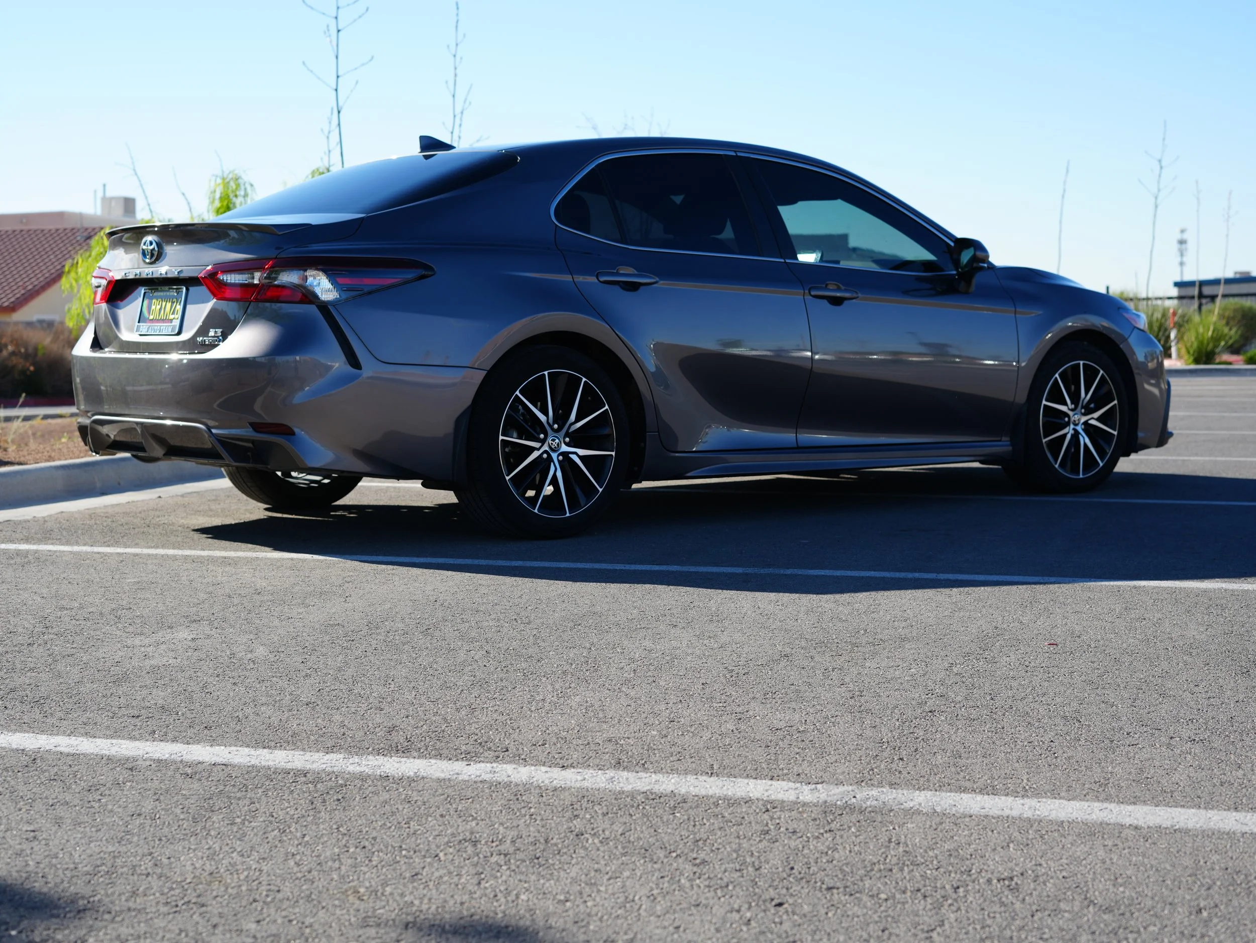 A gray Toyota Camry parked in a parking lot under a clear blue sky with a building and leafless trees in the background.