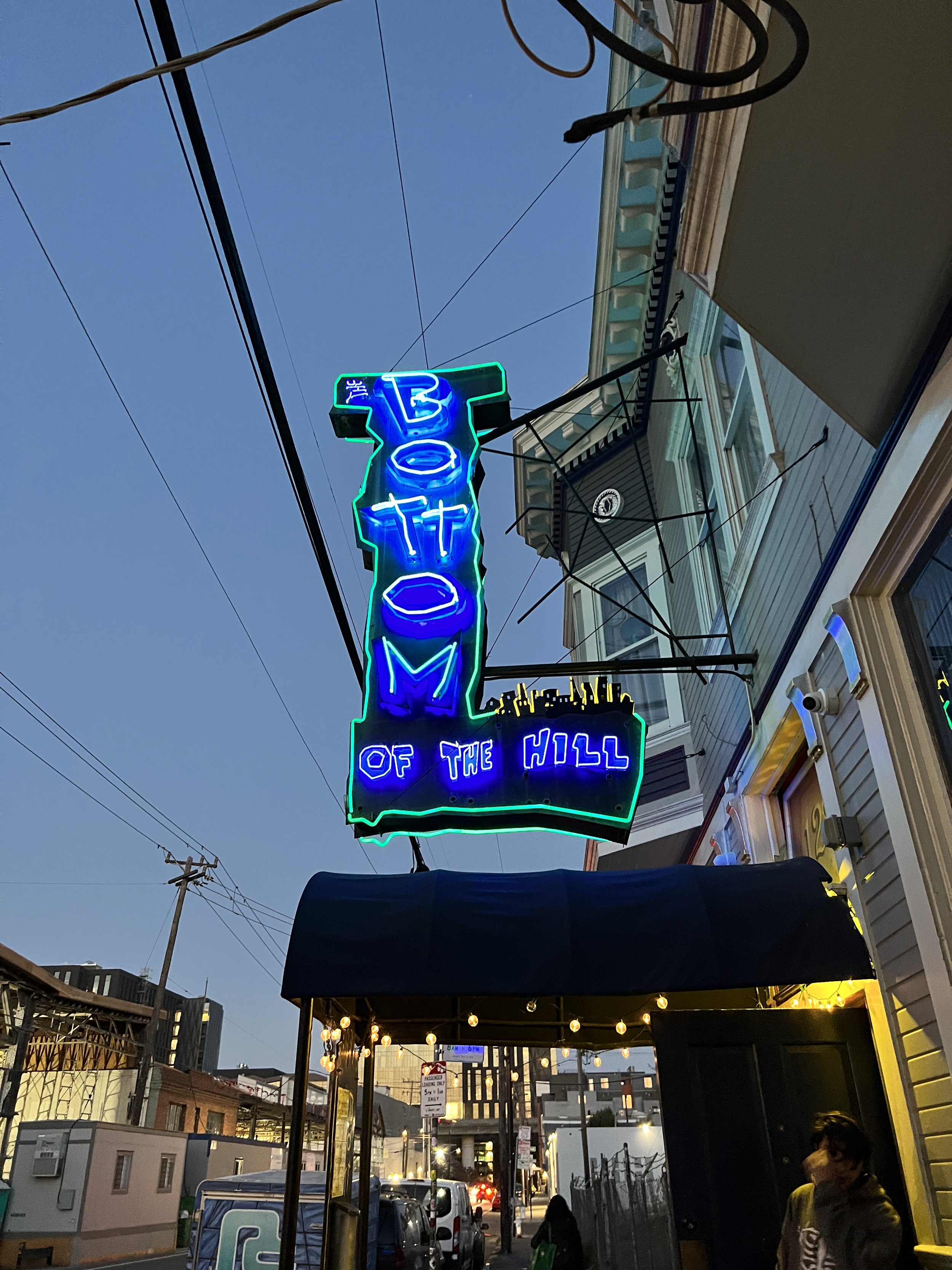 Neon sign reading 'The Bot of the Hill' hanging above a city street with buildings, cars, and people, evening with a darkening sky.