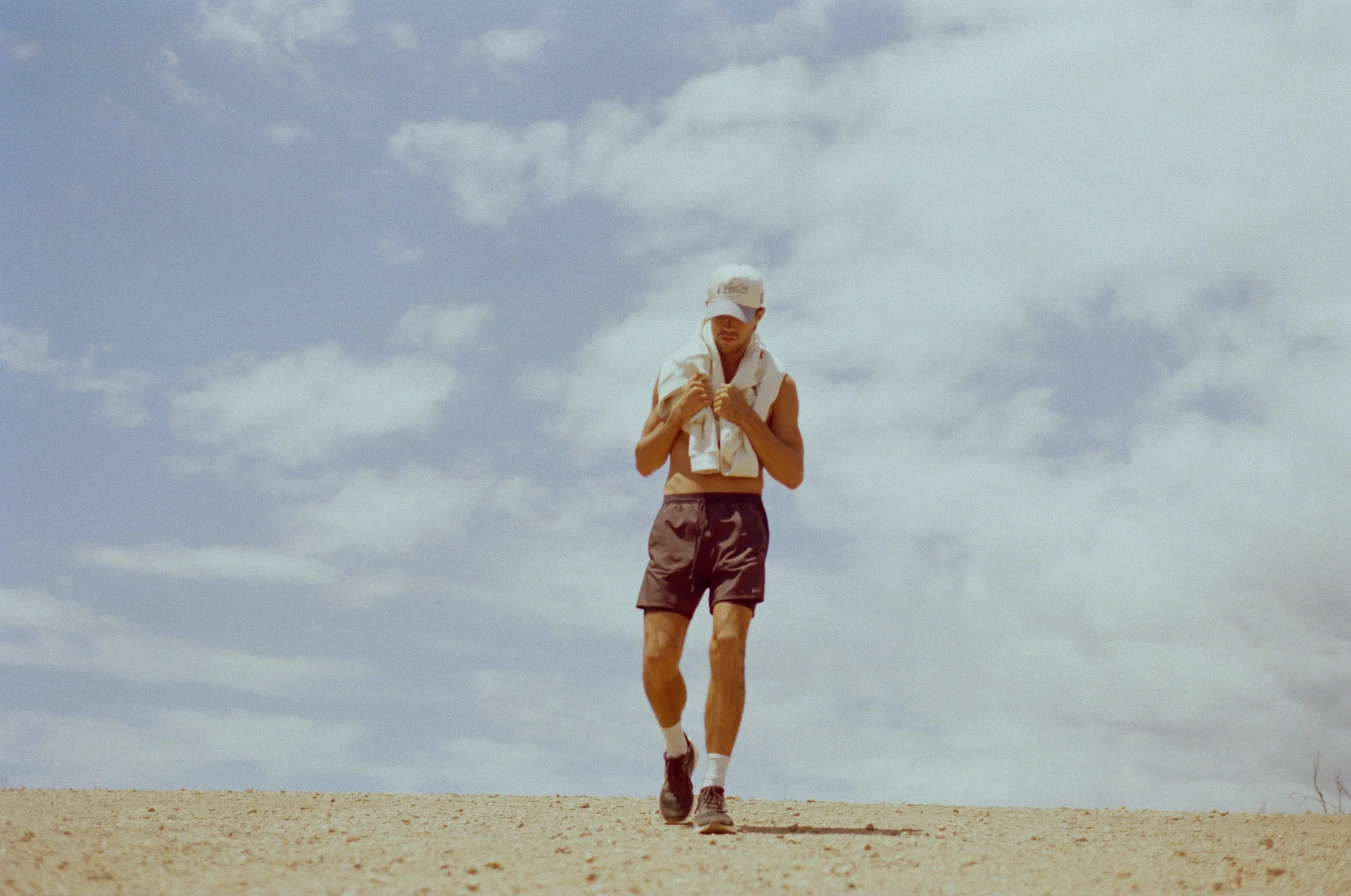 A man is walking outdoors on a sandy terrain, wearing a white cap, sleeveless shirt, shorts, and sneakers. He has a towel around his neck and appears to be in motion, with a cloudy sky in the background.