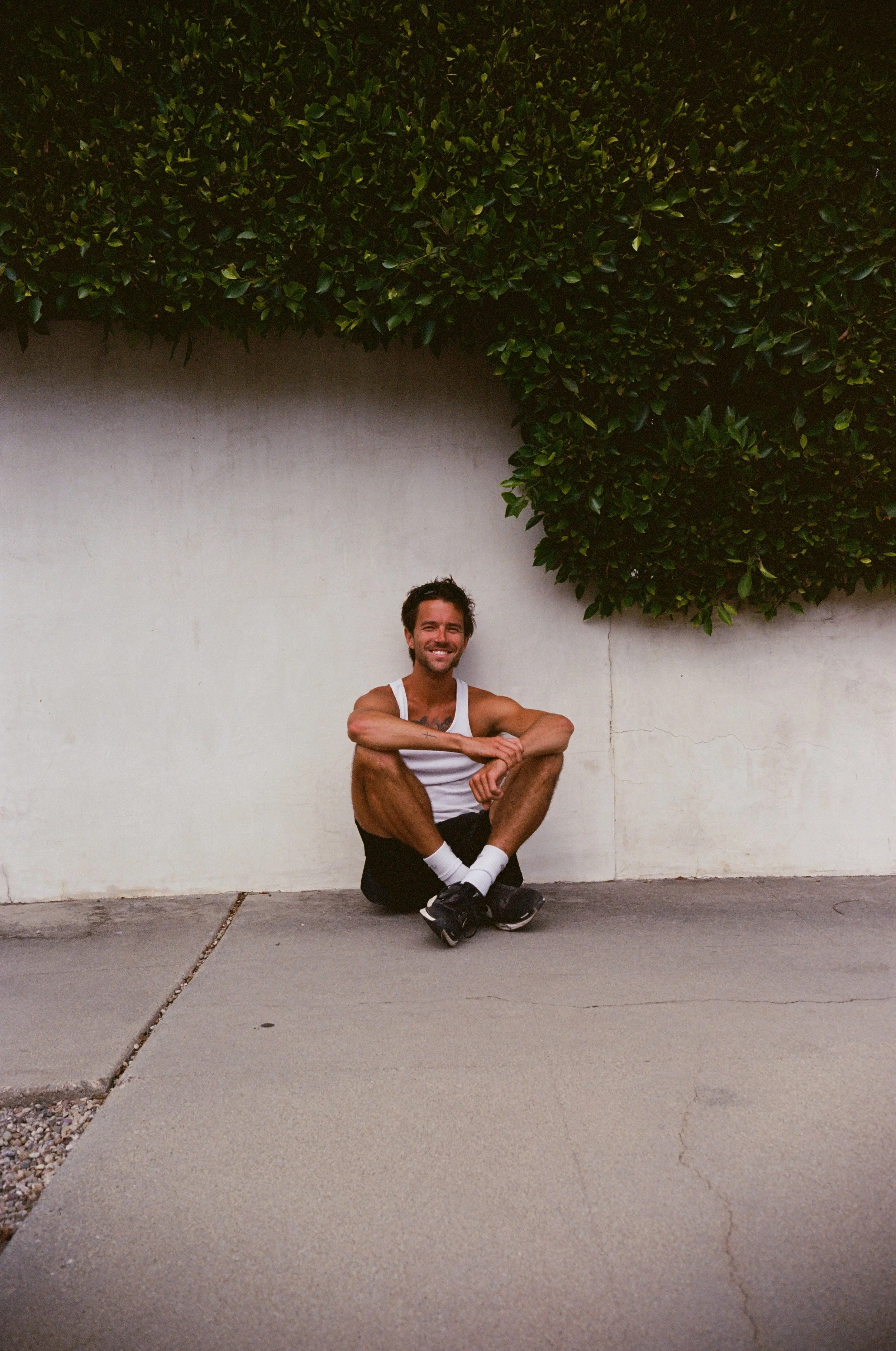 A young man smiling and sitting cross-legged on the sidewalk in front of a white wall with green foliage above.
