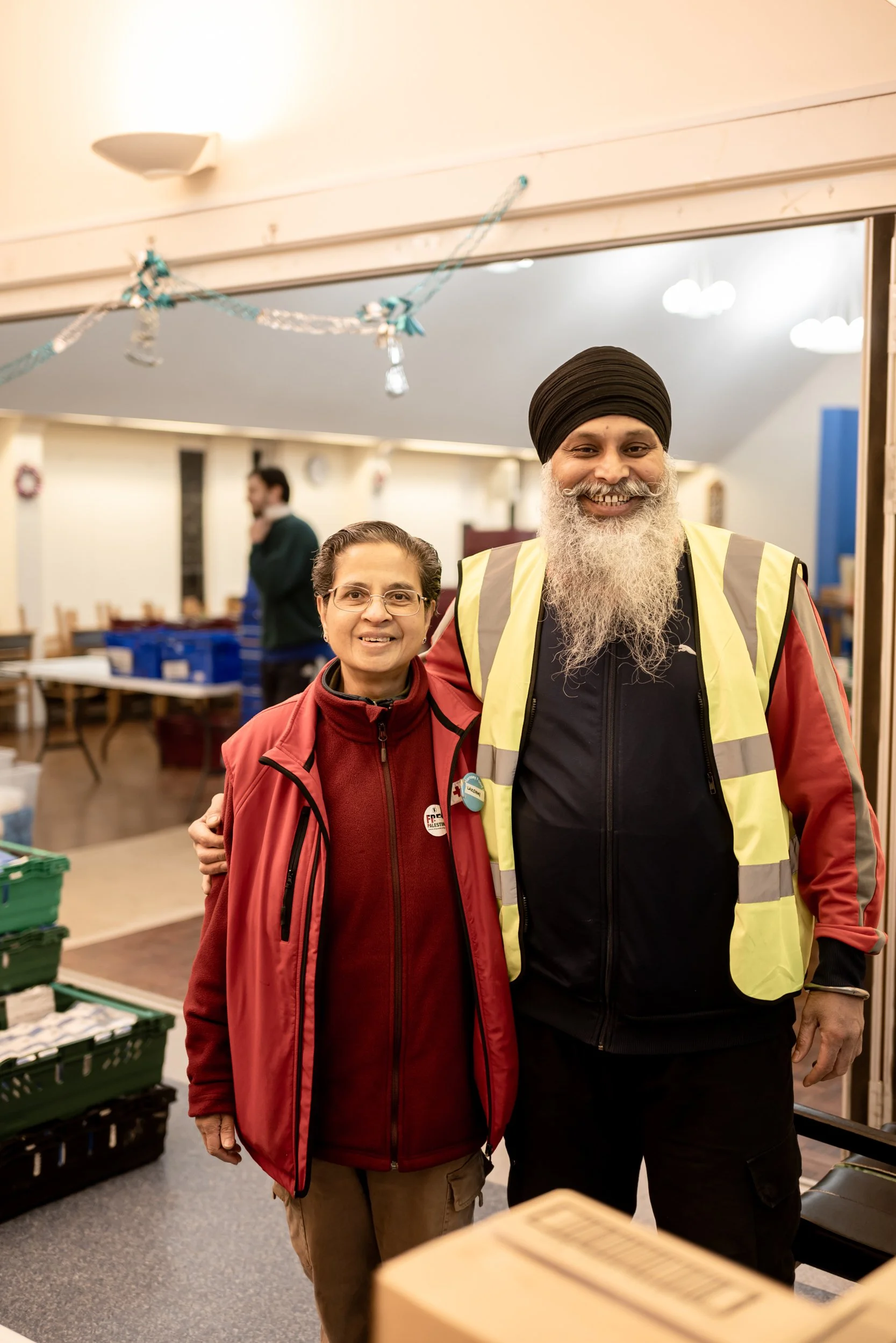 A man and woman smiling inside a community center or volunteer space, standing close with the man’s arm around the woman, with tables and another person in the background.