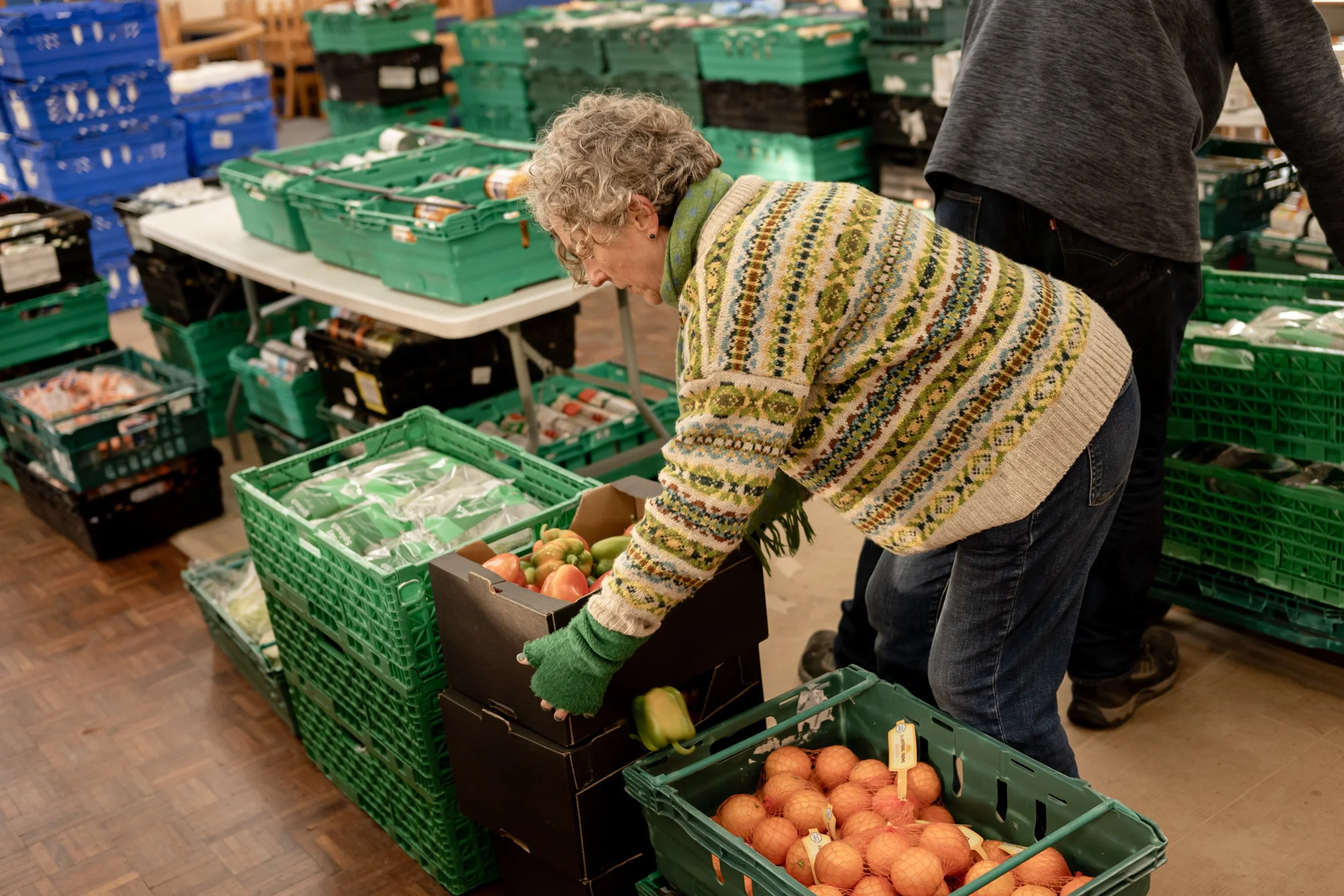 A woman shopping for vegetables at a grocery store, picking up onions from a crate with other produce such as tomatoes and green peppers nearby.