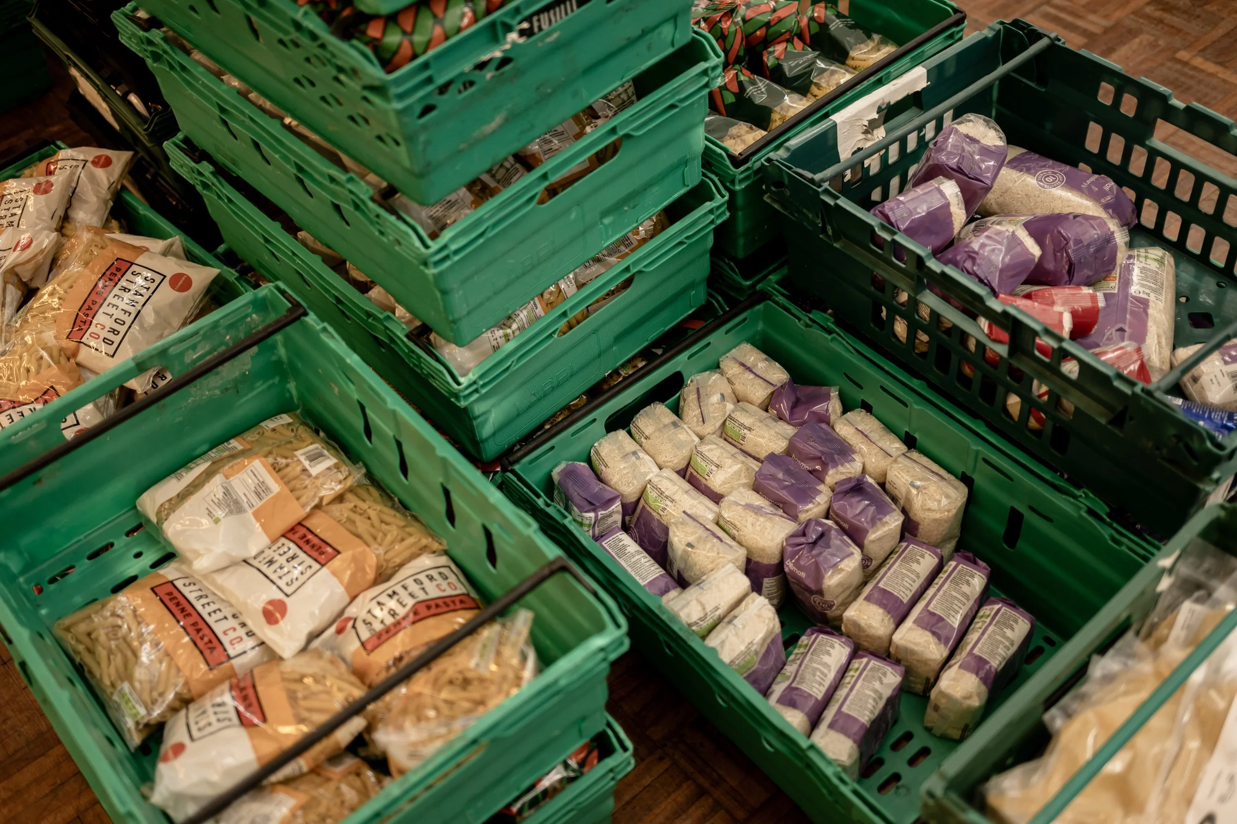 Multiple green plastic crates stacked and filled with various packaged pasta and rice products, arranged in a grocery store or market.