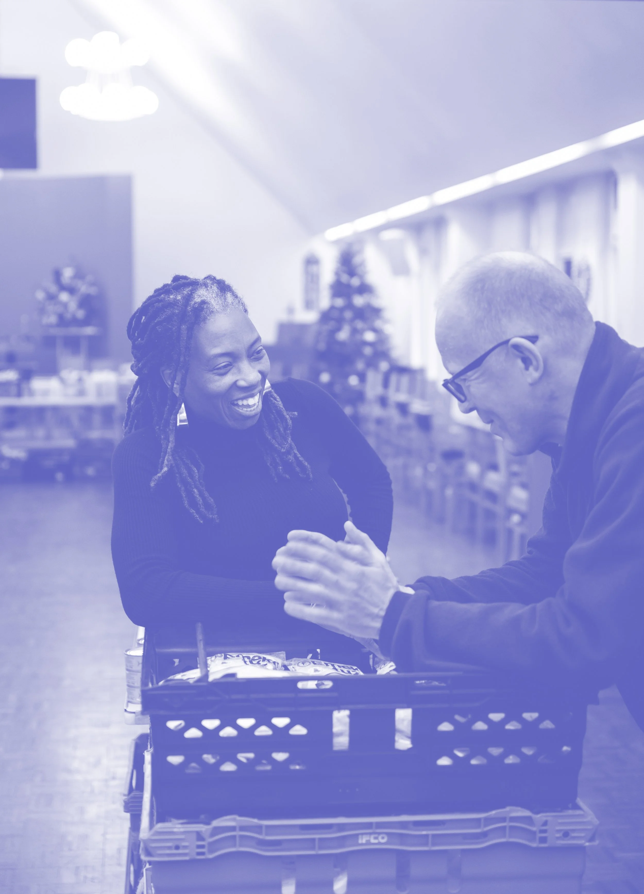 A woman and a man laughing and talking over a crate of groceries in a store decorated for Christmas.