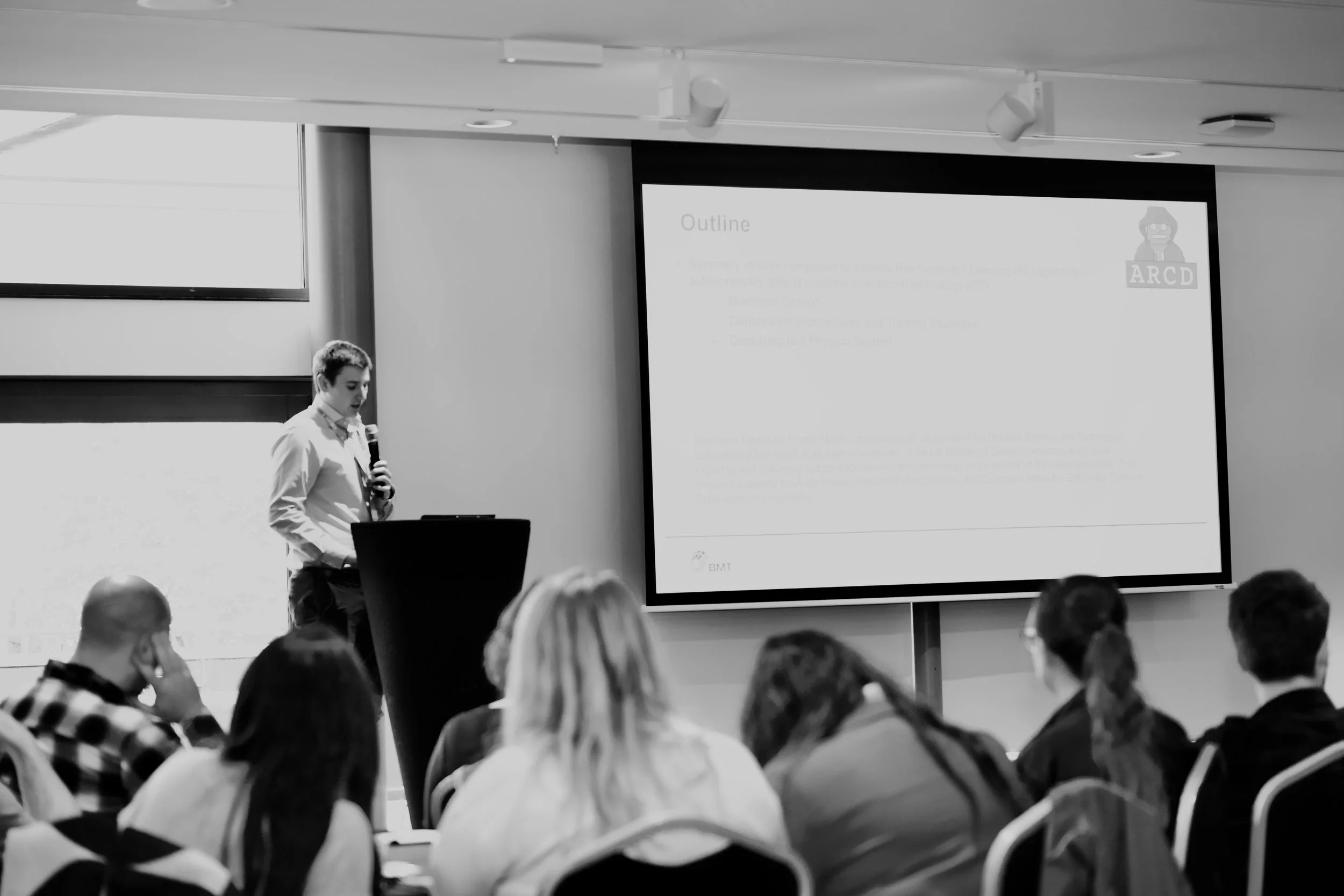 A person standing and speaking into a microphone at a presentation in a conference room with an audience. A large screen behind them displays a slide with the word 'Outline' and some text, including the ARCD logo.