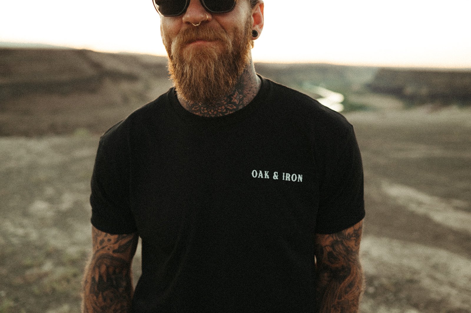Tattooed, bearded man in sunglasses wearing a black Oak & Iron tee in the Idaho high desert at sunset.