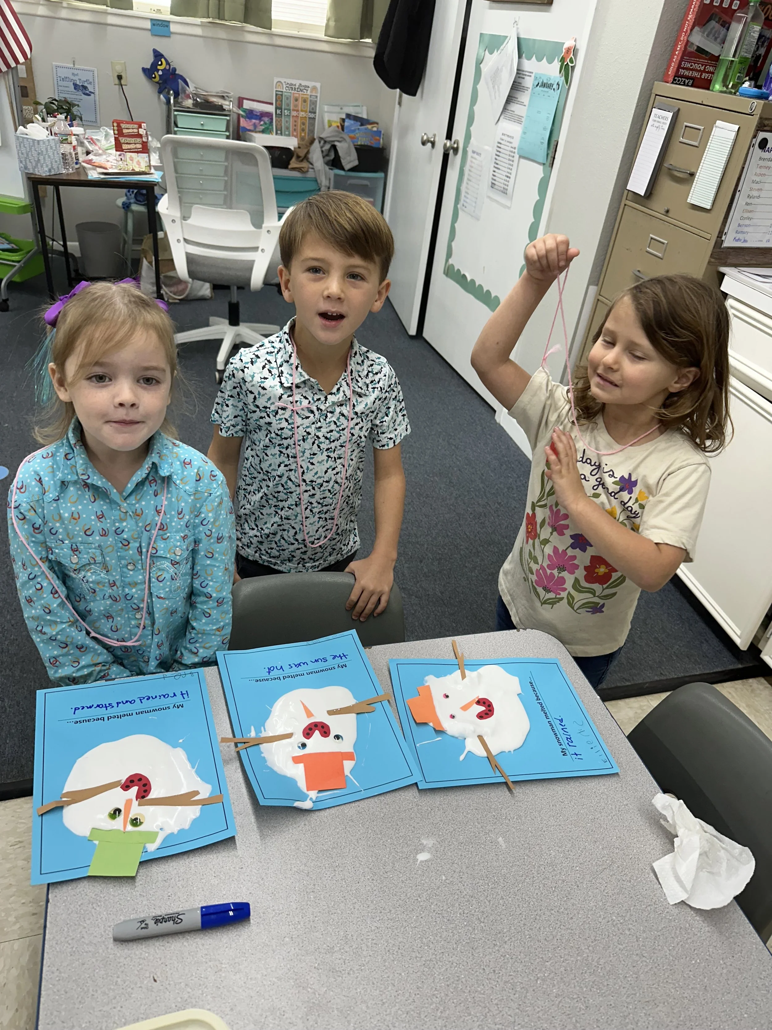 Three children standing around a table with craft projects, including snowman cards decorated with cotton balls, paper accessories, and cotton swabs, in a classroom setting.