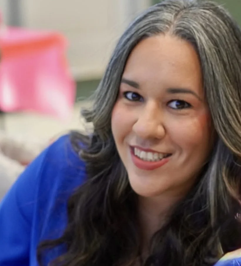 Close-up of a woman with long, wavy gray and brown hair, smiling at the camera. She is wearing a blue top, and the background is out of focus with indistinct objects.