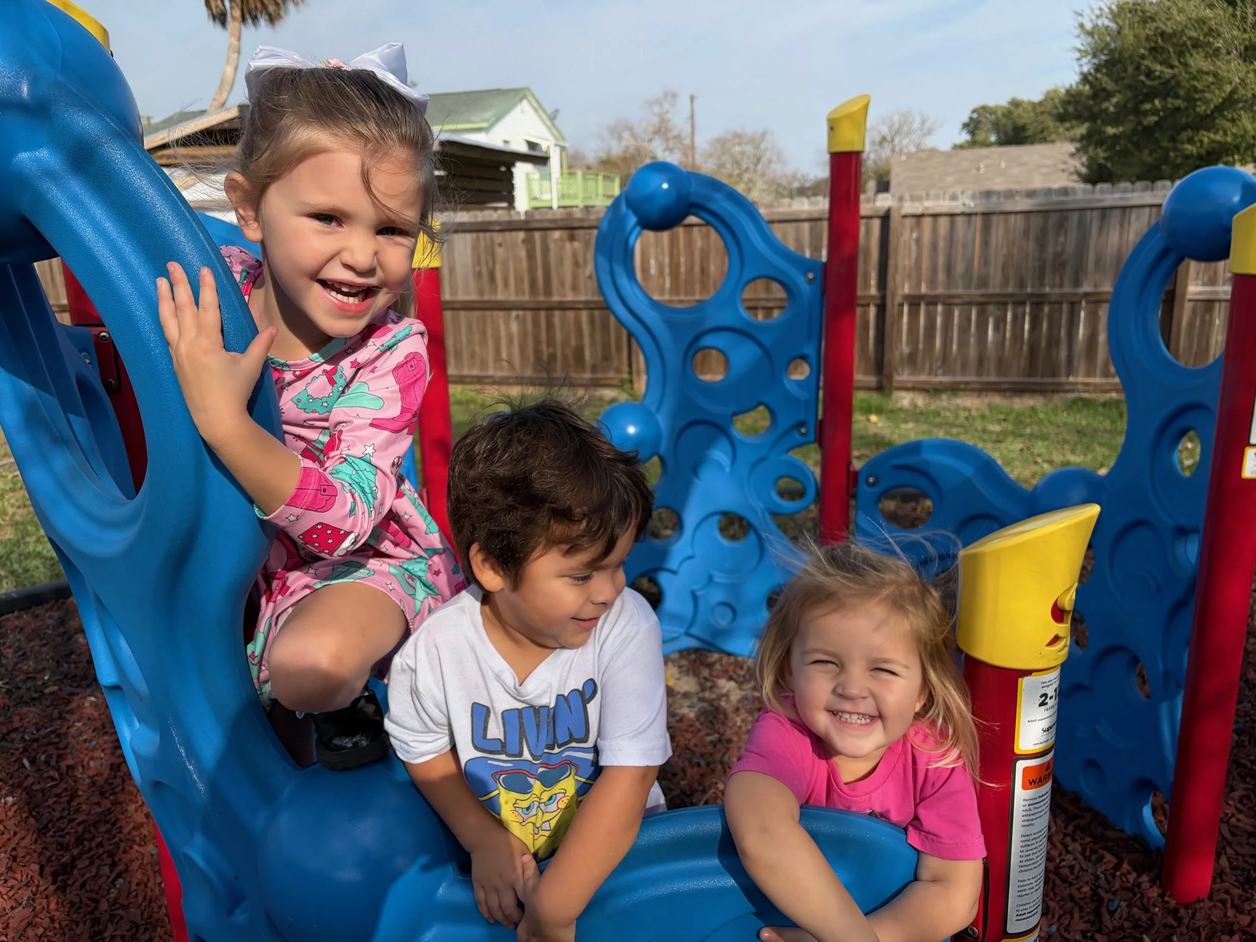 Four children playing on a colorful playground structure outdoors, with a wooden fence and houses in the background. The children are smiling and appear to be having fun.