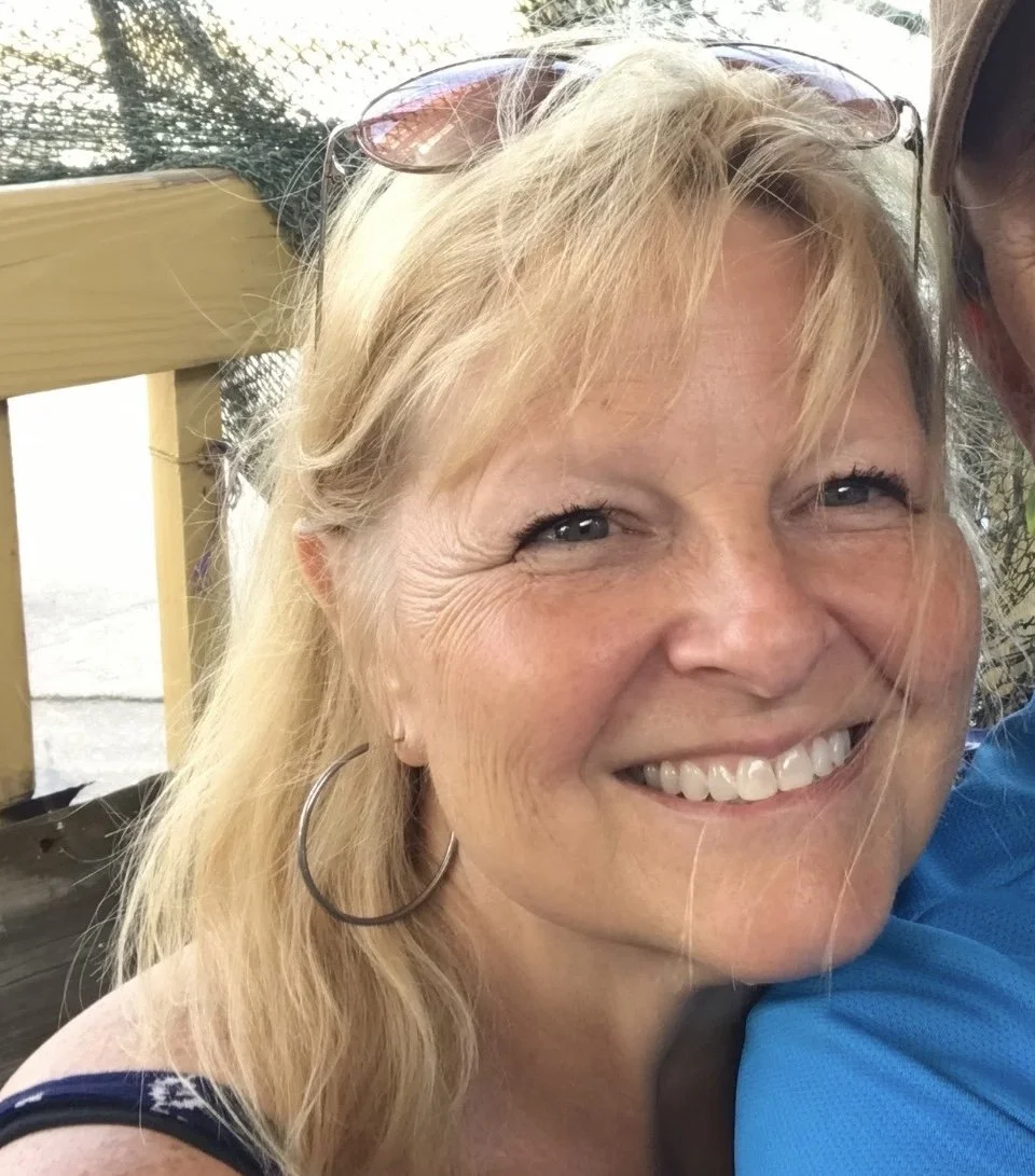 A smiling woman with blonde hair, wearing sunglasses on her head and hoop earrings, leaning close to another person in a blue shirt. They are outdoors, near a wooden railing and trees in the background.
