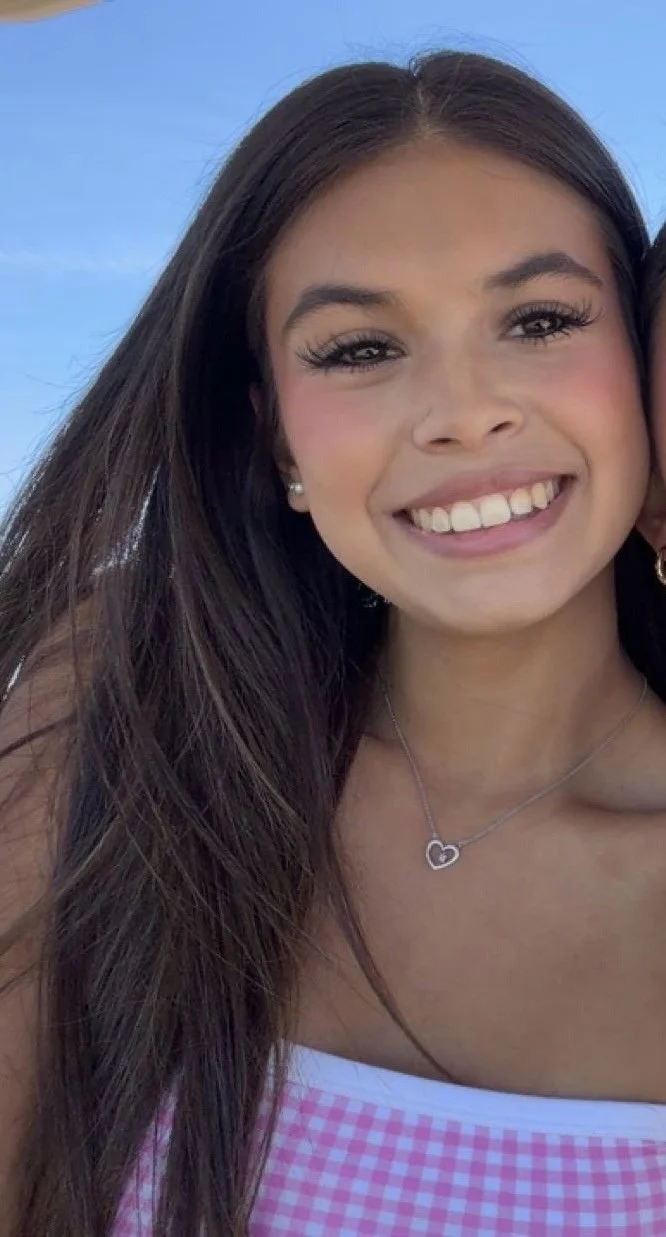 Close-up of a young woman smiling, with long dark hair, wearing a pink and white checkered top, a silver heart-shaped pendant necklace, and pearl earrings, against a blue sky background.