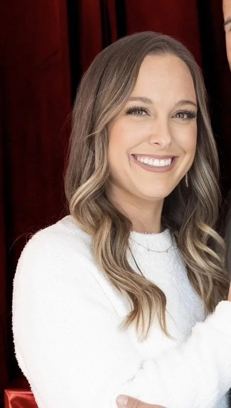 A young woman with long, wavy blonde hair, smiling, wearing a white sweater, standing in front of a red curtain.