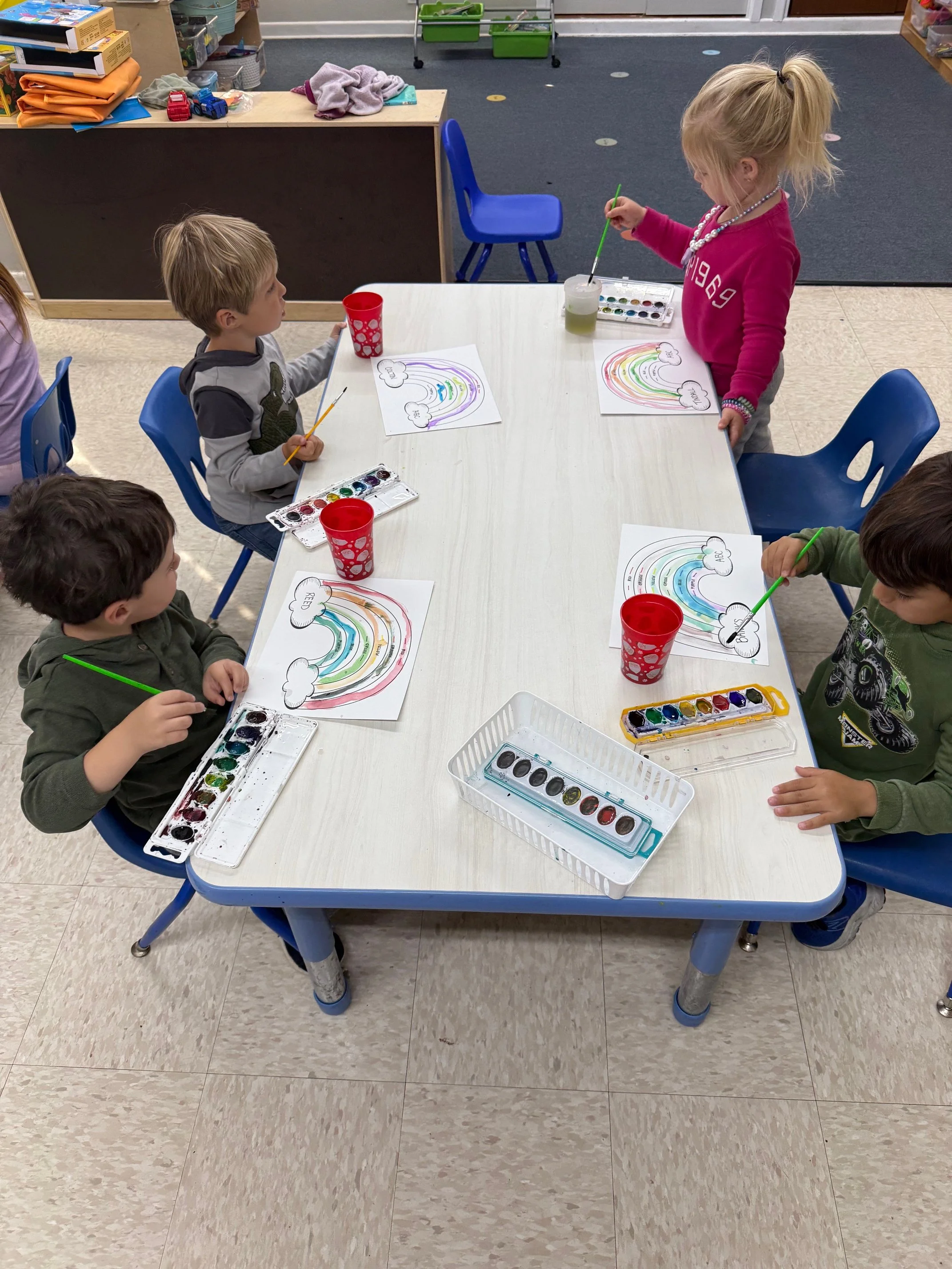 Four young children seated around a table in a classroom, painting rainbows on paper with watercolors.