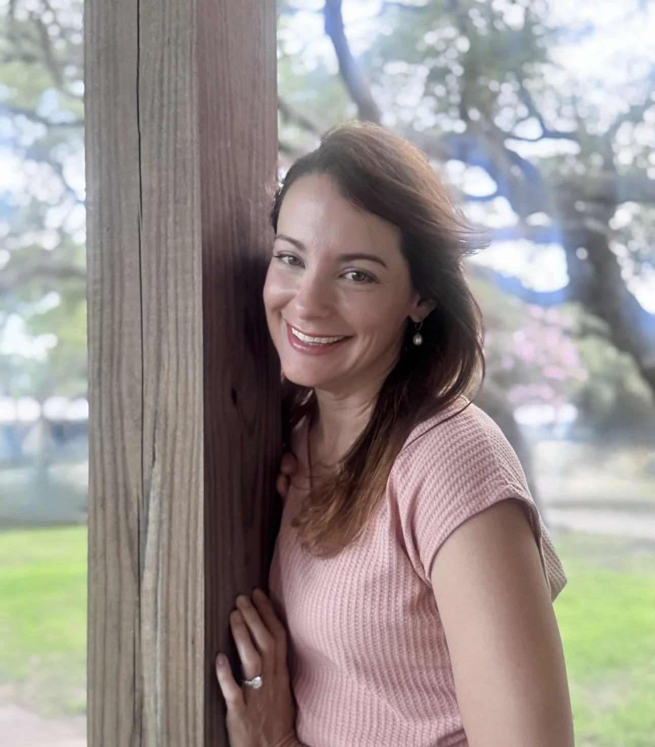 A woman with dark brown hair, smiling and leaning against a wooden post outdoors, with blurred trees and greenery in the background.