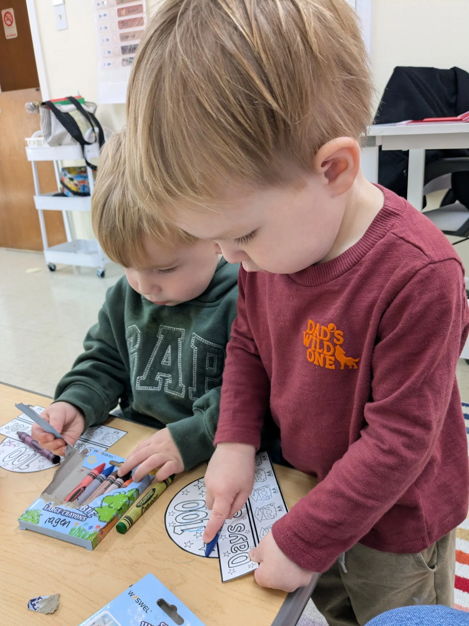 Two young boys are engaged in coloring activity at a desk in a classroom, with crayons and coloring sheets.